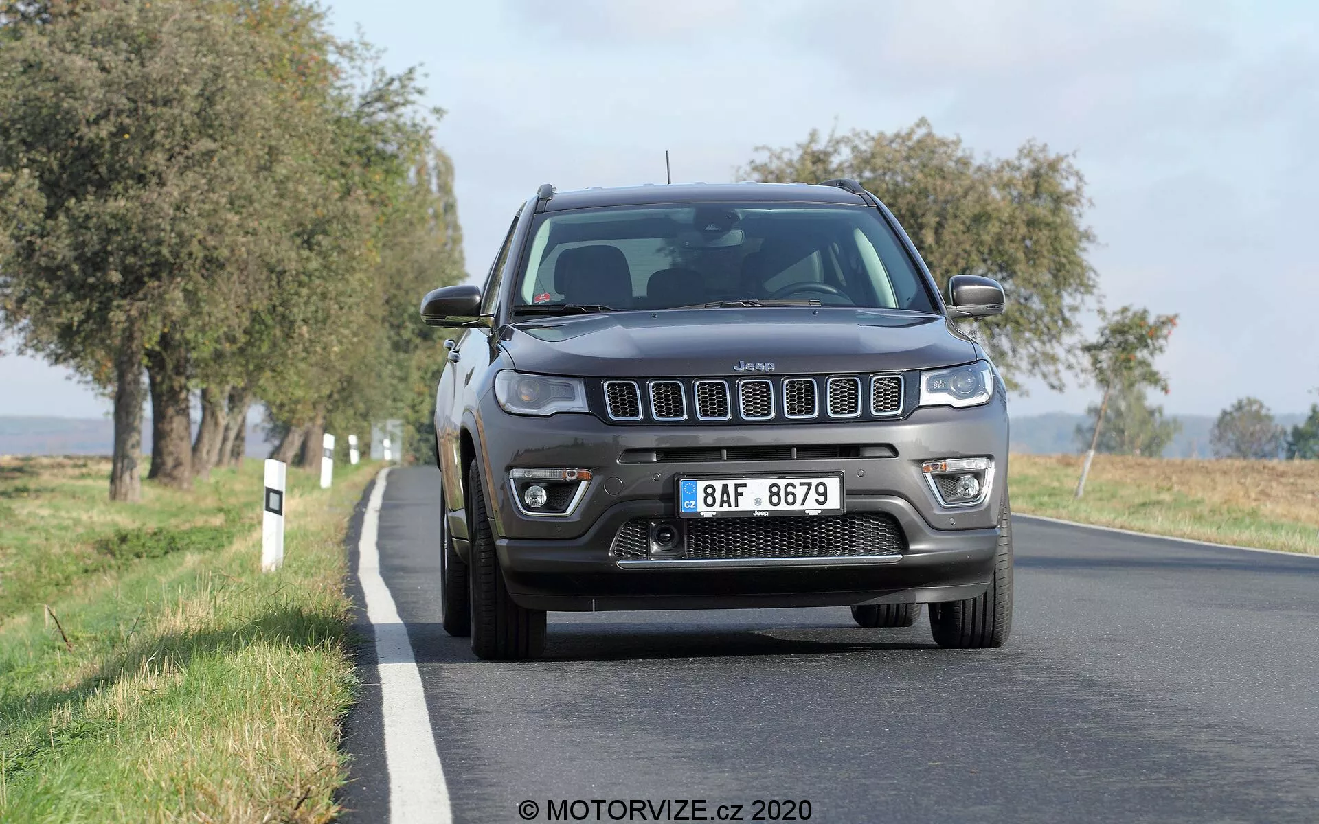 Pogled od spredaj na Jeep Compass 2017, ki vozi po podeželski cesti, obdani z drevesi na obeh straneh. Vozilo ima značilno sedem-režno masko in avtomatske halogenske štirikratne žaromete s funkcijo DRL. Sprednji odbijač vključuje meglenke, na sprednji strani pa je nameščena evropska registrska tablica. Slika zajema vozilo v jasni dnevni svetlobi, poudarjajoč podrobnosti maske, žarometov, pokrova motorja in blatnikov.