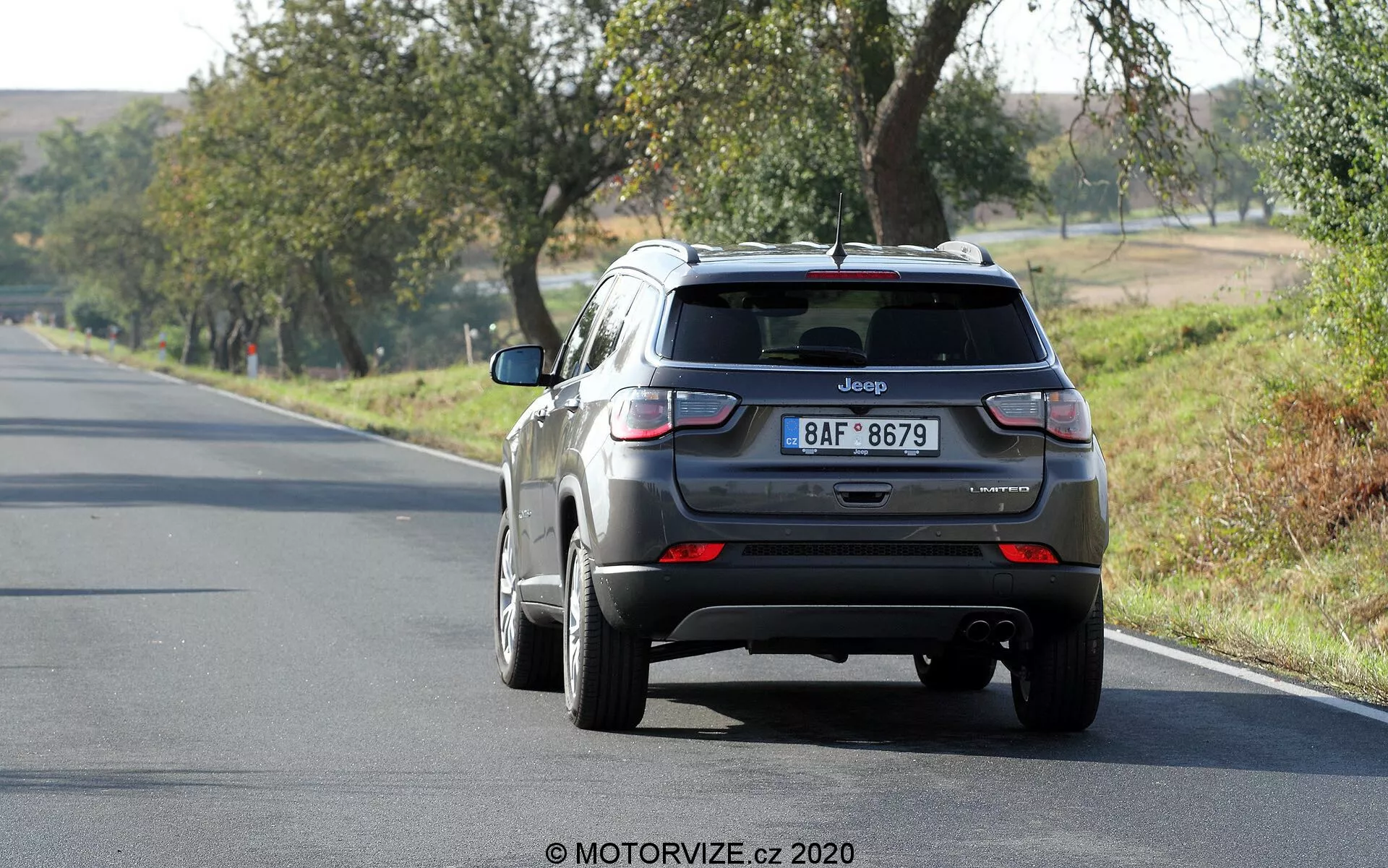 Zadnji pogled na kovinsko sivi Jeep Compass SUV iz leta 2017, ki se oddaljuje po dvopasovni cesti, nameščen nekoliko desno od okvirja. Vozilo na desnem pasu ima vidne lastnosti, kot so zadnja vrata z značko Jeep in registrsko tablico, zadnje vetrobransko steklo z majhnim spojlerjem, zadnje luči z rdečimi in belimi odseki, zadnji odbijač s senzorji in anteno v obliki morskega psa na strehi. Lita platišča so delno vidna. Scena kaže na sončen dan z okoliškimi drevesi in travo, kar nakazuje podeželski ali polpodeželski okolje.