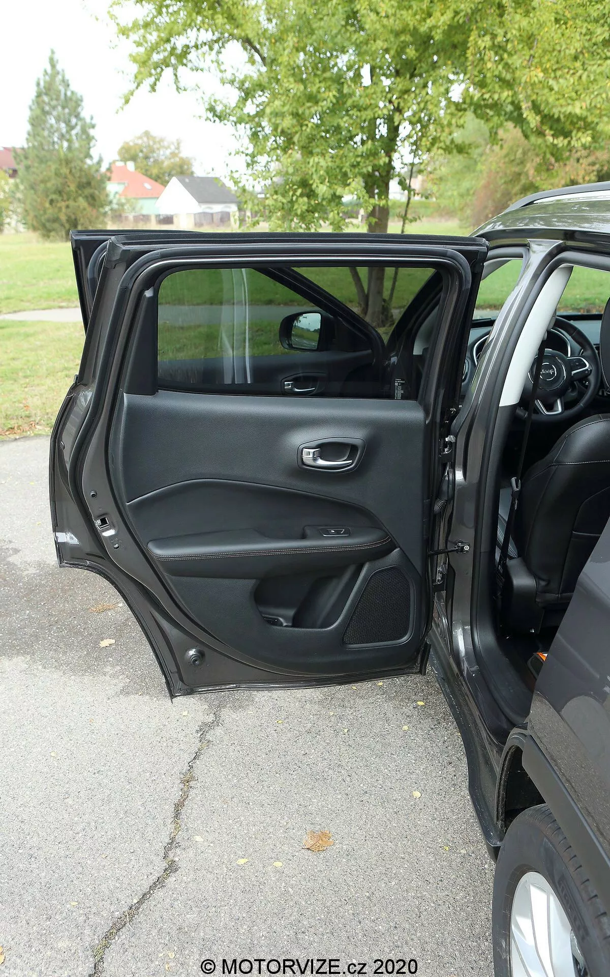 Notranji pogled na Jeep Compass 2017 od zunaj z odprtimi voznikovimi vrati s kovinskim in ergonomsko držalom, odlagalnimi predali, rešetko zvočnika in nadzorno ploščo za upravljanje oken in ključavnice vrat. Vidni so deli armaturne plošče, osrednje konzole in večfunkcijskega volana, kar nakazuje na sodobno notranjost, bogato s funkcijami. Naravna svetloba, oblačno vreme, drevesa v ozadju, ki kažejo na zunanje okolje v parku ali drevoredu.