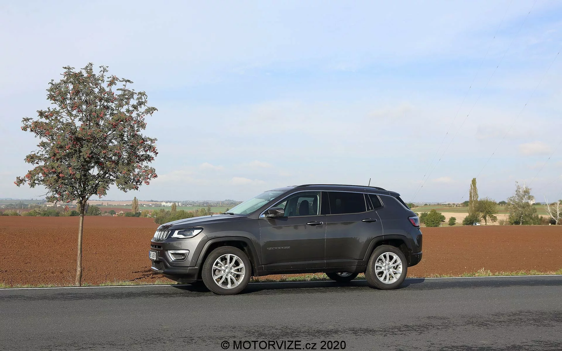 Slika SUV-ja Jeep Compass 2017, parkiranega na ravni asfaltni cesti poleg mladega listnatega drevesa pod jasnim dnevnim nebom. Stranski kot zajema profil vozila od odbijača do odbijača, poudarja njegove sodobne oblikovne elemente, kot so izrazita sprednja rešetka, elegantni žarometi, večkraki aluminijasti platišči, podaljšana stranska ogledala z morebitnimi smerniki in zatemnjena okna. SUV ima dvobarvno barvno shemo in strešni nosilec, s sijajem, ki kaže na kromirane poudarke na sprednji maski. Podeželsko ozadje nakazuje vsestranskost za različne terene. Vodni žig 'MOTORVIZE.cz 2020'.
