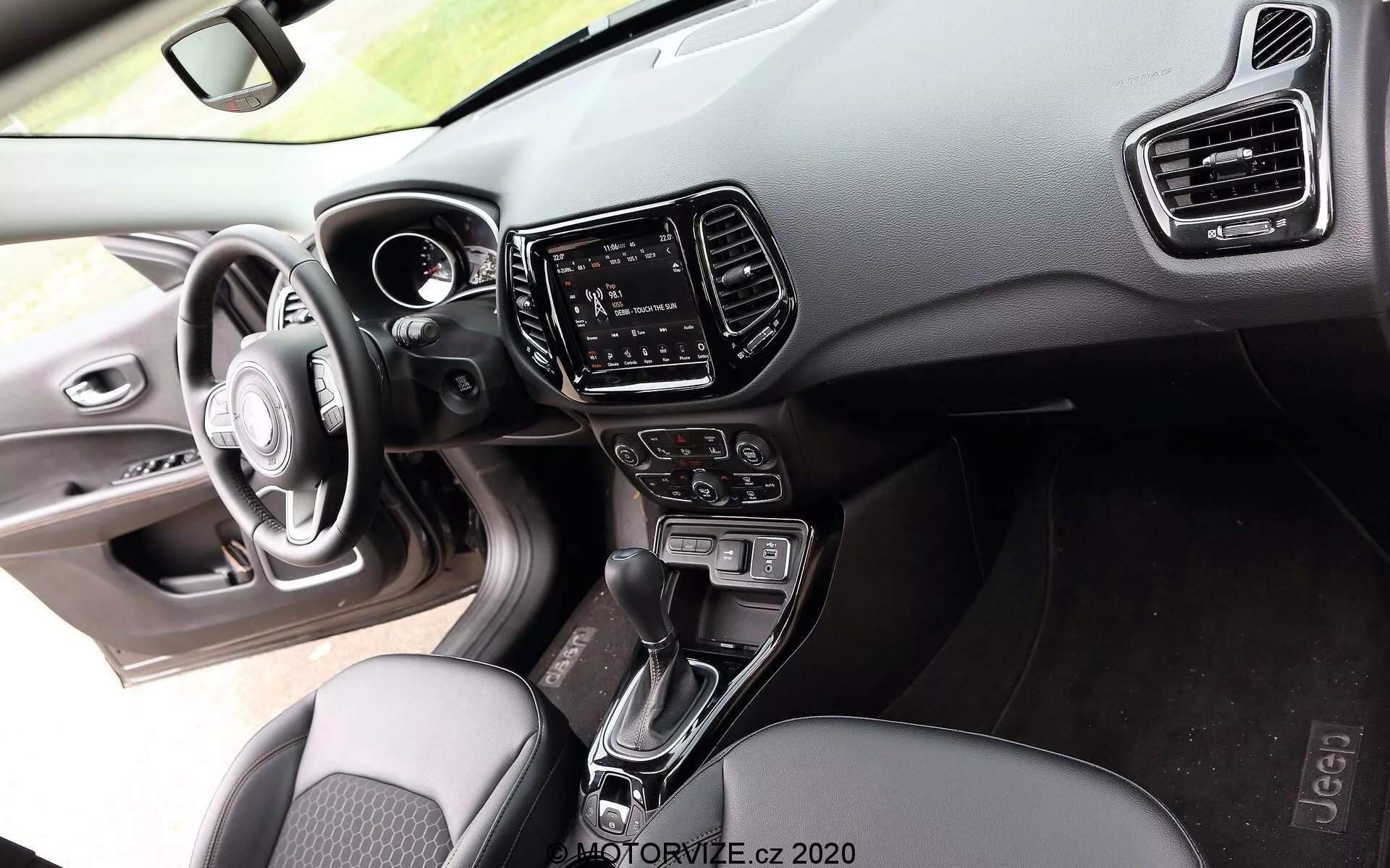 Notranji pogled na Jeep Compass 2017 izperspektive potnika, ki prikazuje trifunkcijski volan z integriranimi kontrolnimi gumbi, delno viden digitalni inštrumentni sklop na armaturni plošči iz mehkih materialov in trde plastike z vidnimi šivi, sistem infotainment s zaslonom na dotik v sredinski konzoli, obdan z navpičnimi klimatskimi odprtinami, avtomatski prestavni ročici med sprednjimi sedeži, gumbe in ročke za nadzor podnebja, tekstilne sedeže z vzorcem in pogled na voznikov vratni panel z nadzornimi funkcijami za nastavljanje oken in vzvratnih ogledal, poudarjajoč udobje in tehnologijo srednjega in visoko-end trga.