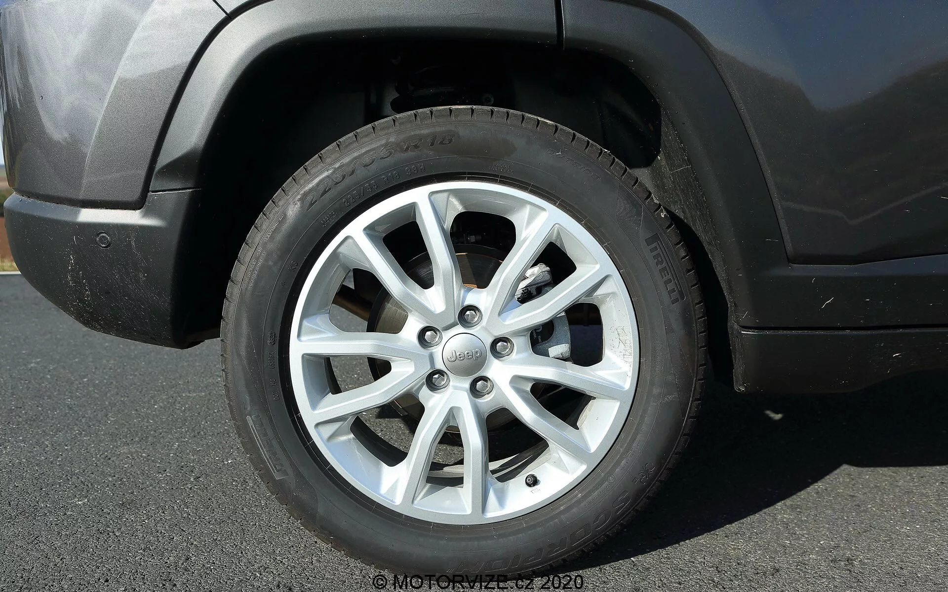 Pogled od blizu na prednje kolo na voznikovi strani 2017 Jeep Compass, s 10-parno večkrako platišče in srebrno obdelavo. Na sredinskem pokrovu je prikazan logotip Jeep. Kolo in pnevmatika kažeta zmerno globino profila in manjše oznake umazanije, kar nakazuje nedavno uporabo. Fotografija je posneta iz nizkega stranskega kota, prikazuje del blatnika kolesa, prednji odbijač in asfaltno površino pod njim. Naravna sončna svetloba in jasne sence kažejo na svetel dan.