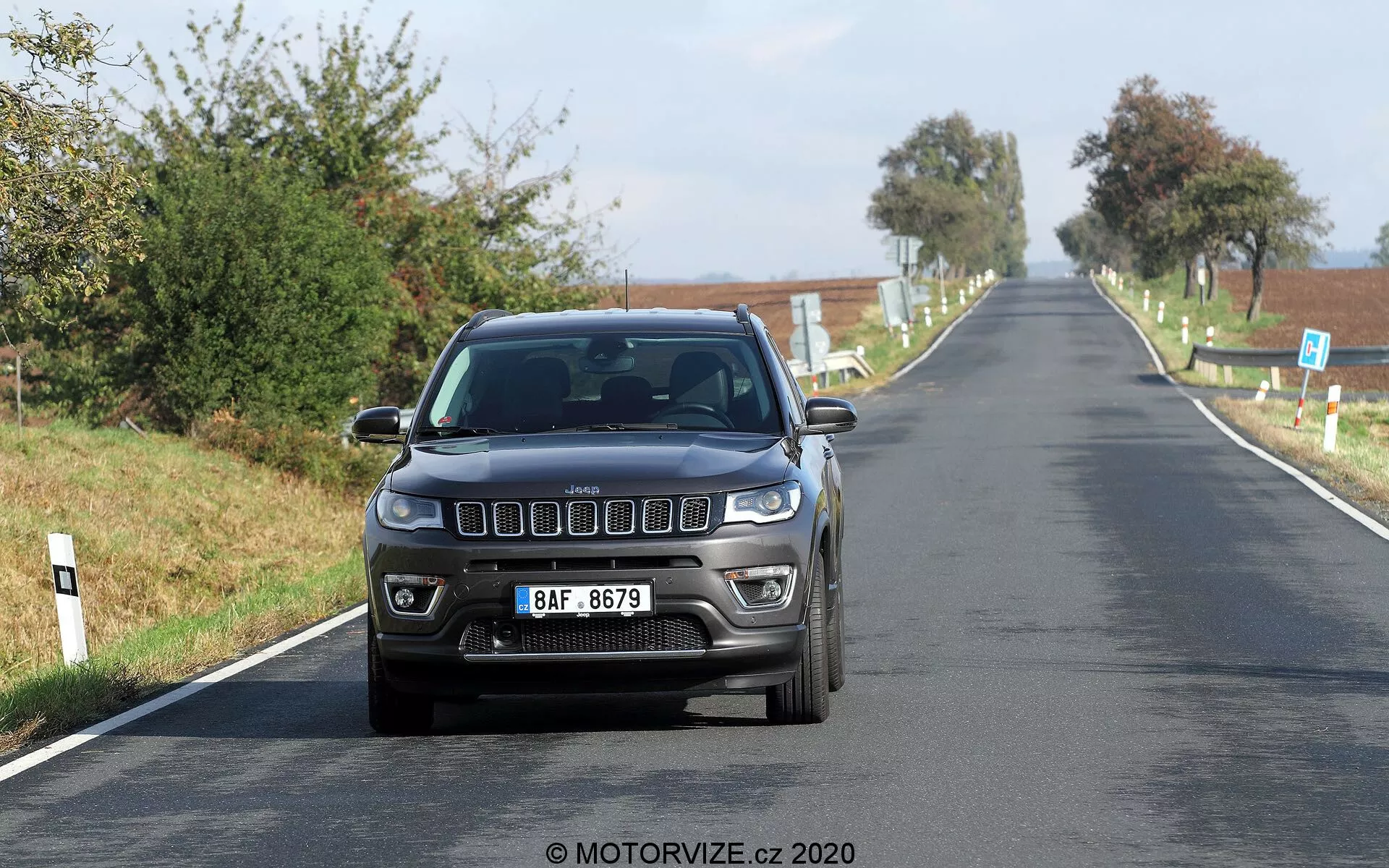 Sprednji pogled na Jeep Compass iz leta 2017, ki z zmerno hitrostjo vozi po podeželski cesti. Slika zajame značilne sprednje lastnosti, vključno z ikonično sprednjo masko s sedmimi režami, modernimi pravokotnimi žarometi in meglenkami, vgrajenimi v odbijač. Avtomobil je fotografiran na ravni oči, kar poudarja njegovo zunanjo zasnovo in osvetljevalno tehnologijo v dobrih naravnih dnevnih svetlobnih pogojih. V sprednjem delu je osredotočena evropska registrska tablica, z jasnim podeželskim ozadjem.