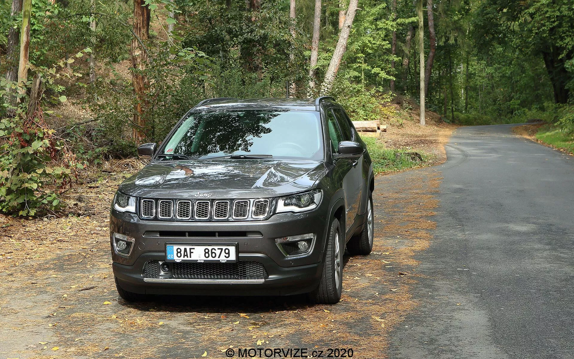 SUV Jeep Compass 2017 na asfaltni cesti obkroženi z zelenjem, obrnjen proti fotoaparatu pod rahlim levim kotom, prikazuje sprednji del in vozniško stran. Vidne značilnosti vključujejo vodoravno rešetko na sprednji strani z osrednjim emblejem, pravokotne LED žaromete, ohišja meglenk v spodnjem odbijaču, konturirano kapuco, ki odseva svetlobo, zrcalne pokrove v barvi karoserije, rahlo zatemnjena okna, večkrako lito kolo in konturiran blatnik. Ozadje vključuje drevesa in listje, kar kaže na gozdnato obcestno območje.