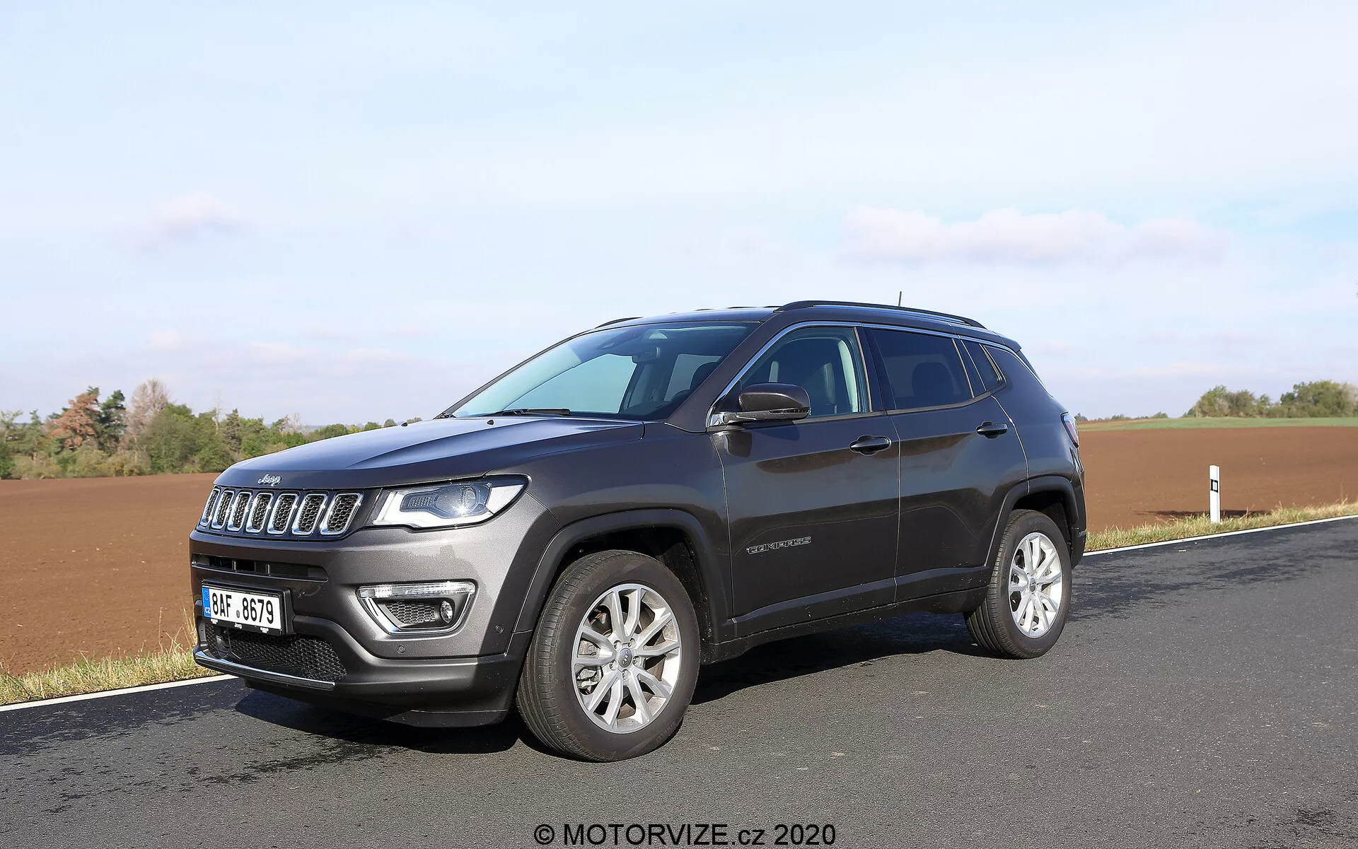 Sodobni SUV parkiran ob cesti s poljem v ozadju, posnet iz kotnega sprednjega kota. Fotografija poudarja sedem rešetkasto prednjo masko SUV-ja, značilne žaromete z LED lučmi, aluminijasta platišča in temno kovinsko barvo. Vidni so stranski detajli, kot so linija oken, ročaji vrat, bočna ogledala z integriranimi smerniki ter rahlo nagnjena linija strehe. Na sprednjih vratih je vidna tudi značk, ki ponuja možne informacije o različici vozila.