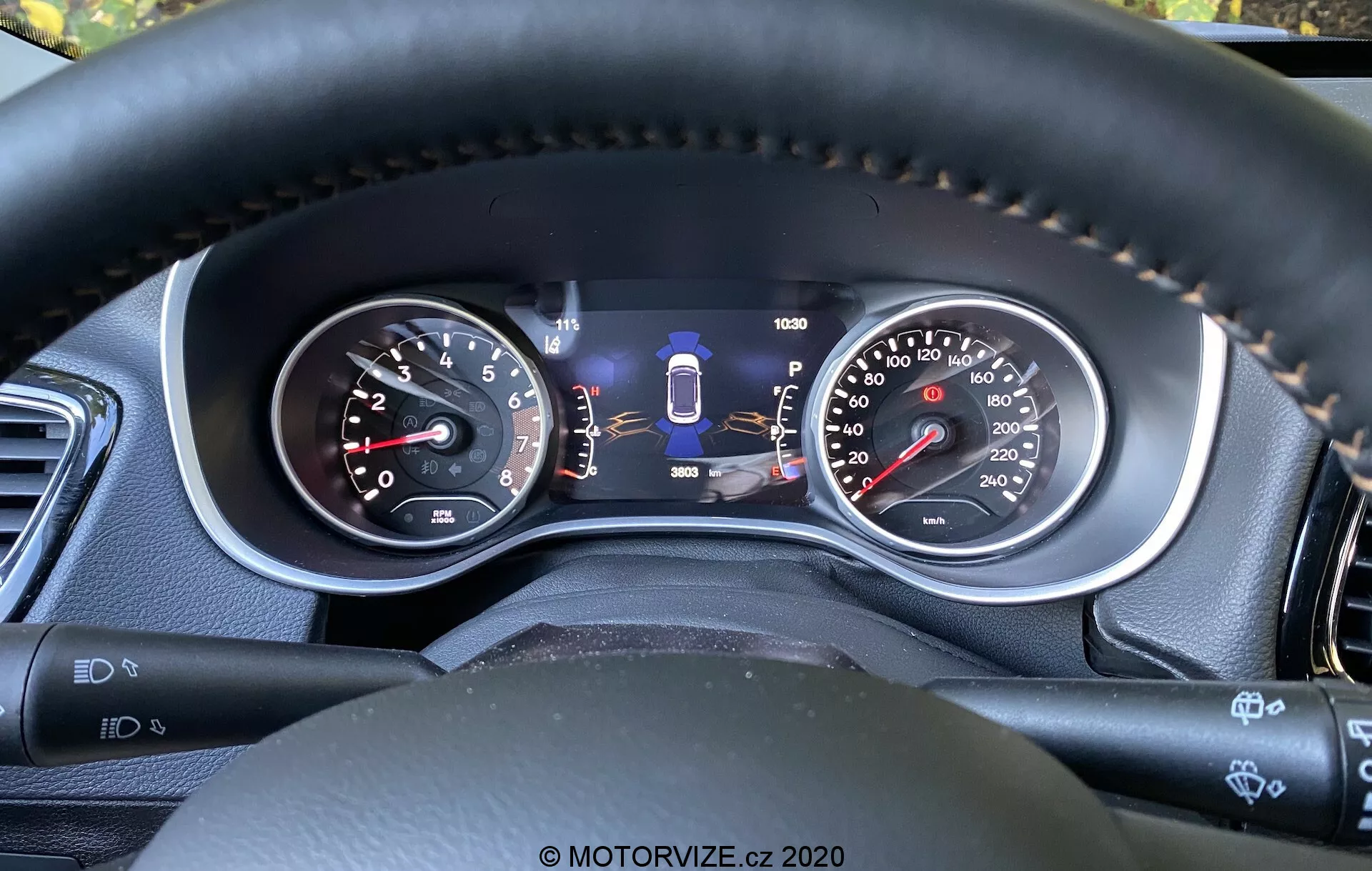Pogled od blizu na instrumentno ploščo Jeep Compass 2017 iz voznikove perspektive, ki prikazuje analogne merilnike in osrednji digitalni zaslon. Značilnosti vključujejo tahometer, dvouhitnostni merilnik hitrosti, indikator izbire prestave, merilnik goriva, števec kilometrov ter dodatne indikatorje varnostne tehnologije, kot je pomoč pri ohranjanju voznega pasu. Šivanje na volanu iz materiala, podobnega usnju, poudarja kakovostne materiale in udobje, medtem ko nadzorni ročaji in gumbi nakazujejo napredne funkcije udobja in pomoči vozniku.