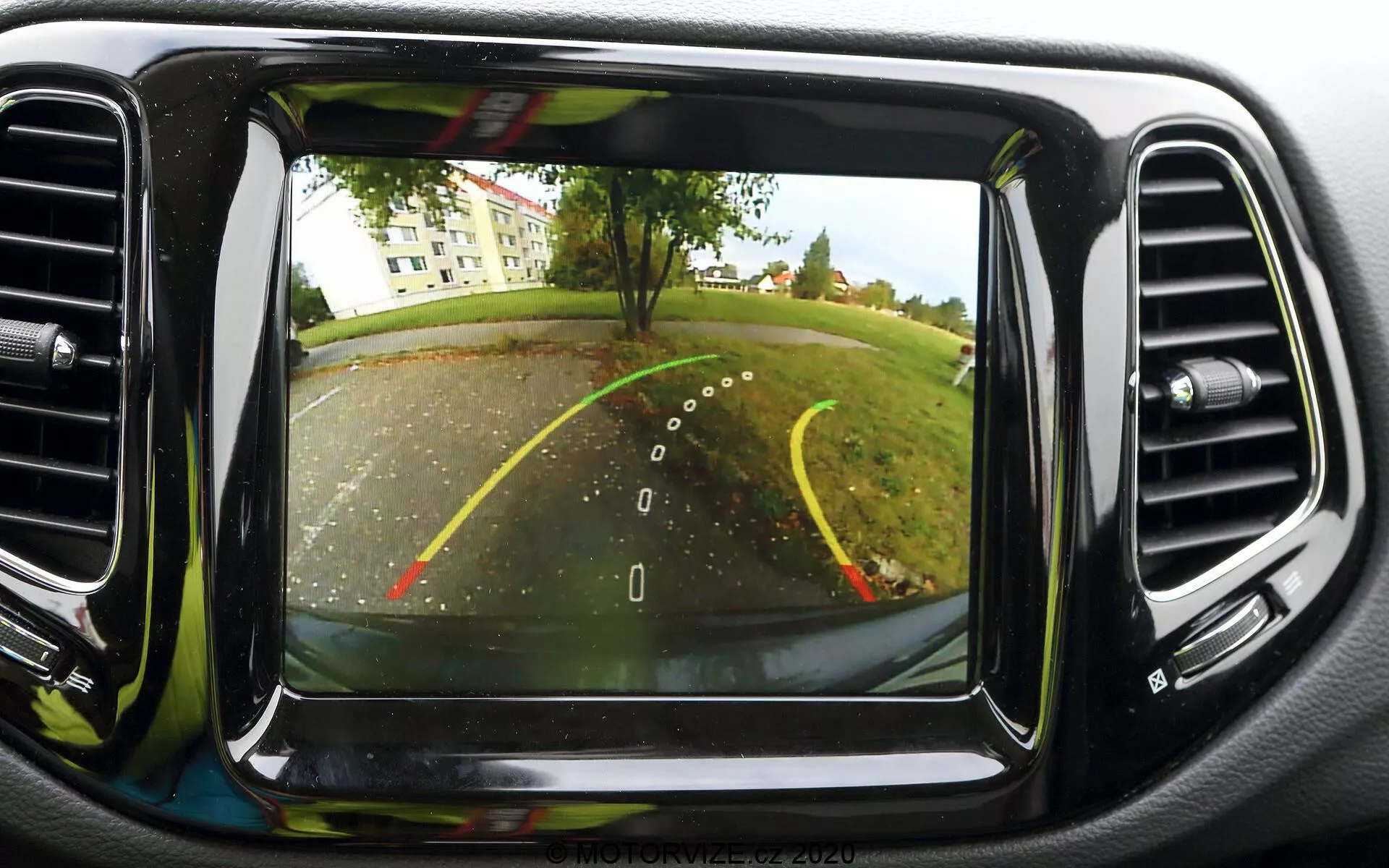 Slika sistema vzvratne parkirne kamere na armaturni plošči vozila Jeep Compass 2017. Kamera, nameščena zadaj pri registrskih tablicah, zagotavlja širok kot za pogled za vozilom. Vidne so prekrivne smernice za vzvratno vožnjo, ki označujejo širino vozila in predvideno pot s pomočjo rumenih in rdečih črt za razdaljo. Prikazana scena vključuje parkirišče ali dovozno pot z drevesi in travnatim območjem v ozadju. Zaslon je osrednje nameščen na armaturni plošči, obdan z odprtinami za klimatsko napravo, in ima čisto kvaliteto slike, značilno za sodobne avtomobilske tehnologije.