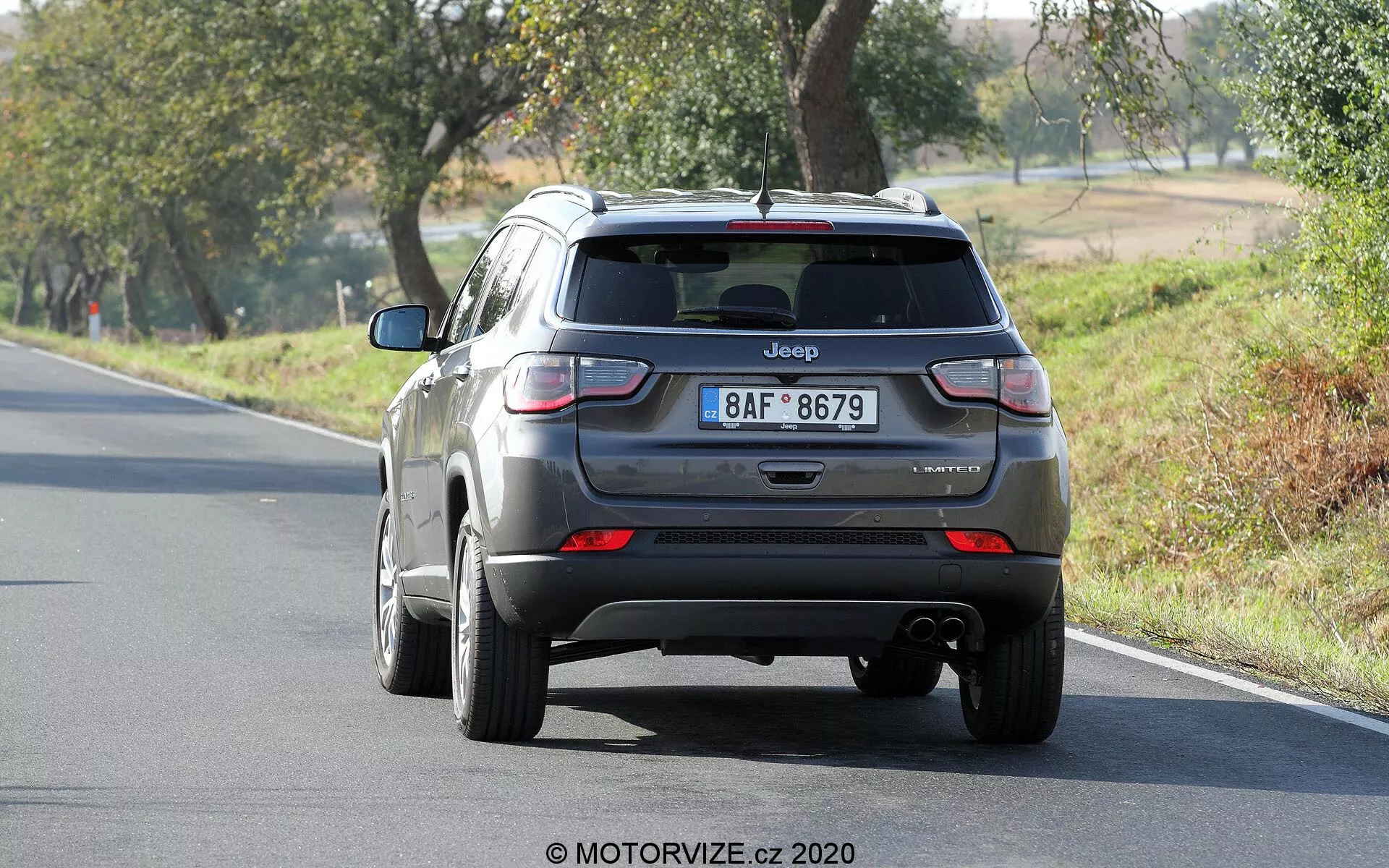 Zadnji pogled na srebrni Jeep Compass 2017, ki prikazuje zadnja vrata, zadnji odbijač in zadnje luči. Vidna sta napis 'Jeep' in ime modela 'Compass', z nejasno berljivo oznako nivoja opreme. Slika jasno prikazuje zadnje luči, izpušno cev pod odbijačem in avtomobil, postavljen na dvopasovno cesto, obdano z drevesi v podeželskem okolju.