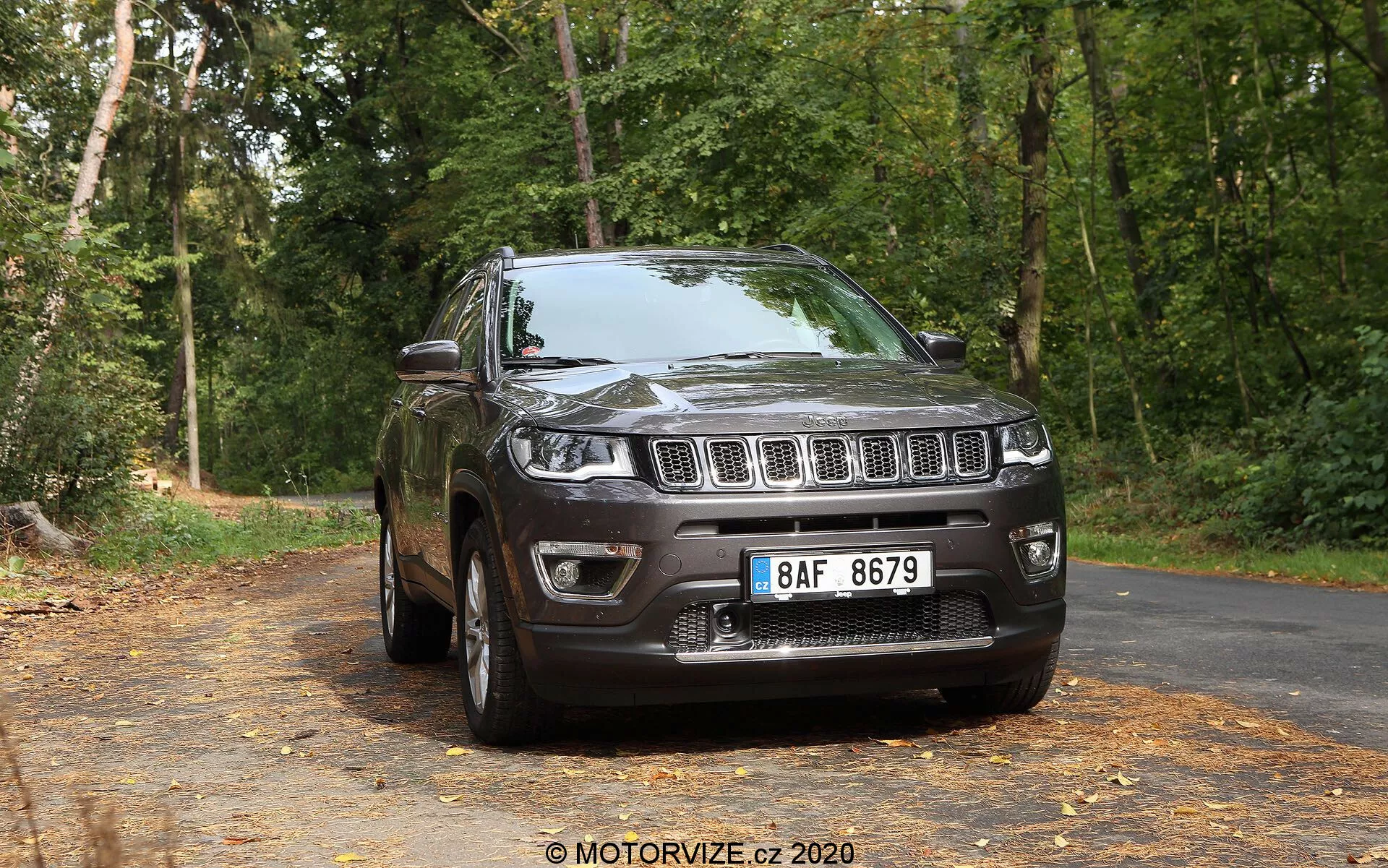 Pogled na prednji tričetrtinski del temno obarvanega SUV Jeep Compass 2017 na mirni, z listjem pokriti asfaltni cesti z ozadjem dreves v gozdnem okolju. Slika prikazuje sprednjo rešetko z značilnimi navpičnimi režami, obdano s žarometi, ki segajo v blatnike, dobro definiran sprednji odbijač z meglenkami in oblikovan pokrov motorja. Stranski profil razkriva zlitinska platišča, viden blatnik, stranska ogledala, kljuke vrat, obrise oken in strešni sistem, vse to osvetljeno z naravno svetlobo.