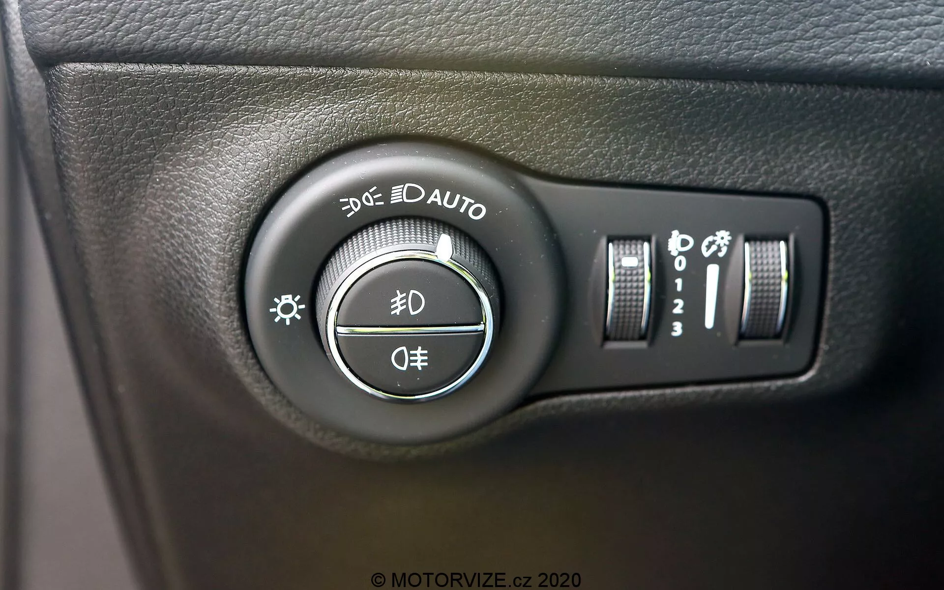 Pogled od blizu na stikalo za nadzor žarometov pri Jeep Compass 2017, ki se nahaja na armaturni plošči na levi strani volanskega obroča. Slika, posneta pod rahlim kotom, poudarja vrtljivo stikalo z nastavitvami za parkirne luči, dnevne luči, avtomatske žaromete, zasenčene in dolge luči ter položaj izklopa. Dodatni nadzorni elementi na desni vključujejo navpični drsnik za svetlost armaturne plošče in gumb, ki lahko kaže na samodejne dolge luči ali sistem izravnave žarometov. Stikalo je nameščeno na površino iz teksturirane črne plastike, z jasnimi belimi oznakami in rahlo sijočimi odsevi.