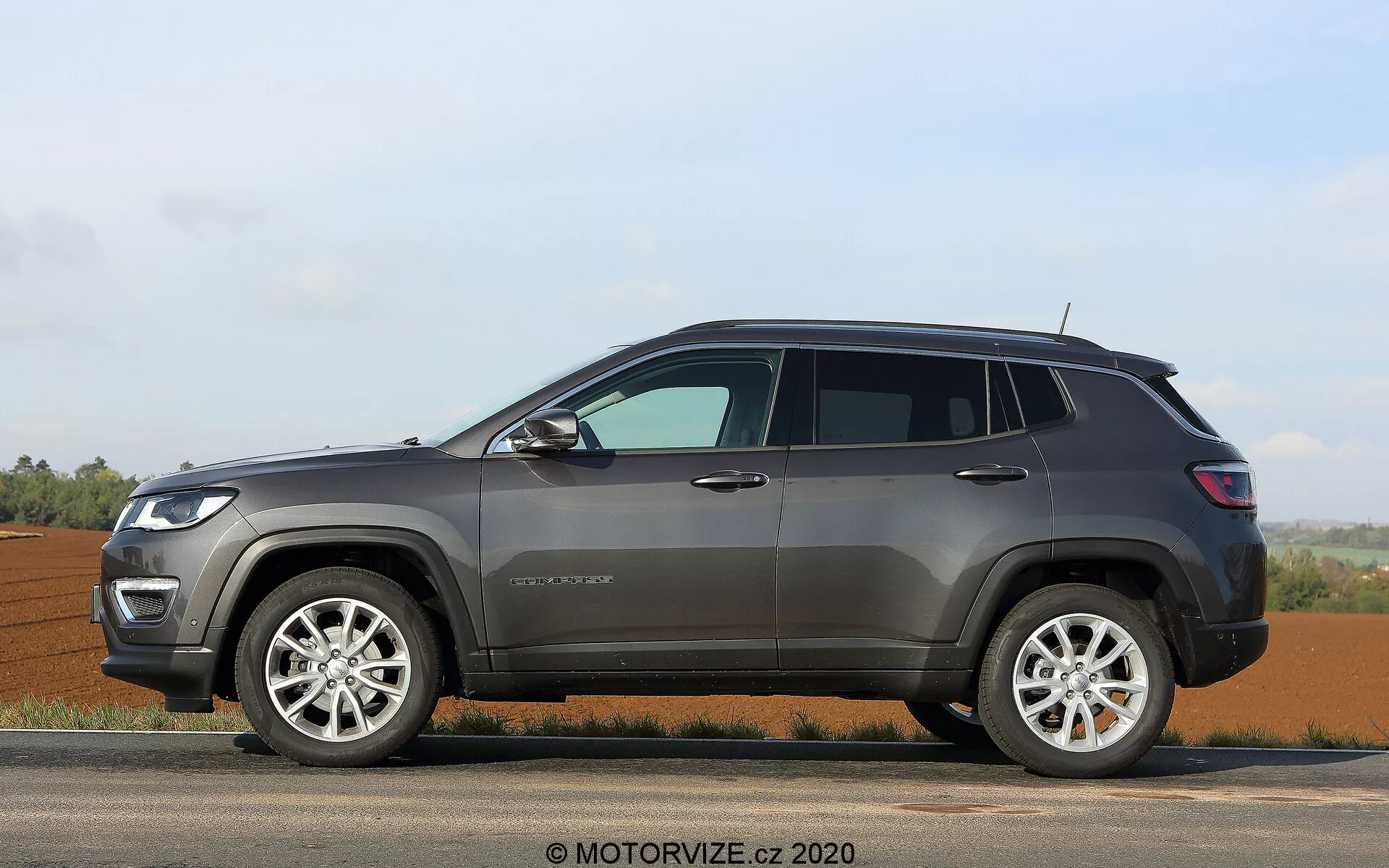 Stranski profil sodobnega SUV Jeep Compass 2017, parkiranega na ravni površini, prikazuje potniško stran z dvignjenimi okni, štirimi vrati, vidnim B-stebričkom in četrtinskim oknom na območju C-stebrička. Očitna so lita kolesa z večkrakim dizajnom in pnevmatike z izrazitimi stranskimi stenami, kar kaže na možnost uporabe na cesti in izven nje. Zunanjost ima plastično oblogo okoli kolutnih lokov in spodnjega dela karoserije, strešne nosilce za dodatni tovor, verjetno z vgrajenimi smerokazi in robustne konture vrat. Na sprednjem blatniku je badge, ki označuje model ali raven opreme, in povišan položaj vozila nakazuje visoko odmik od tal, primeren za različne terene. Statičen, proti ruralnemu ozadju z odprtimi polji in jasnim obzorjem, pod blagimi svetlobnimi pogoji.