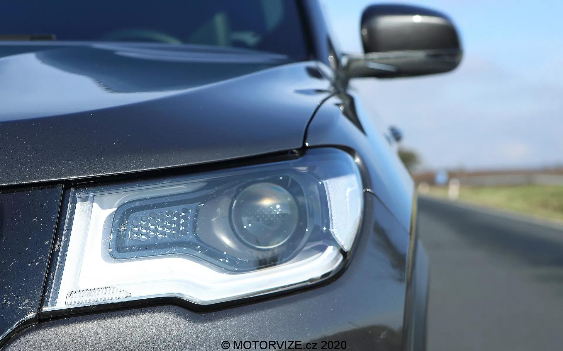 Odpoved v bližini prednjega levega dela Jeep Compass 2017, osredotočena na sklop žarometov z nizkega kota na tleh. Sodobni žaromet vključuje projektorski snop in LED dnevne luči, razporejene v značilnem vzorcu. Ohišje žarometa, elegantno, se gladko povezuje s temno obarvano karoserijo, ki odseva jasno modro nebo. Delno vidno stransko ogledalo se ujema z barvo karoserije in lahko vključuje stranski kazalnik. Plitva globina polja poudarja podrobnosti žarometa in sprednje maske proti zamegljenemu ozadju ceste.