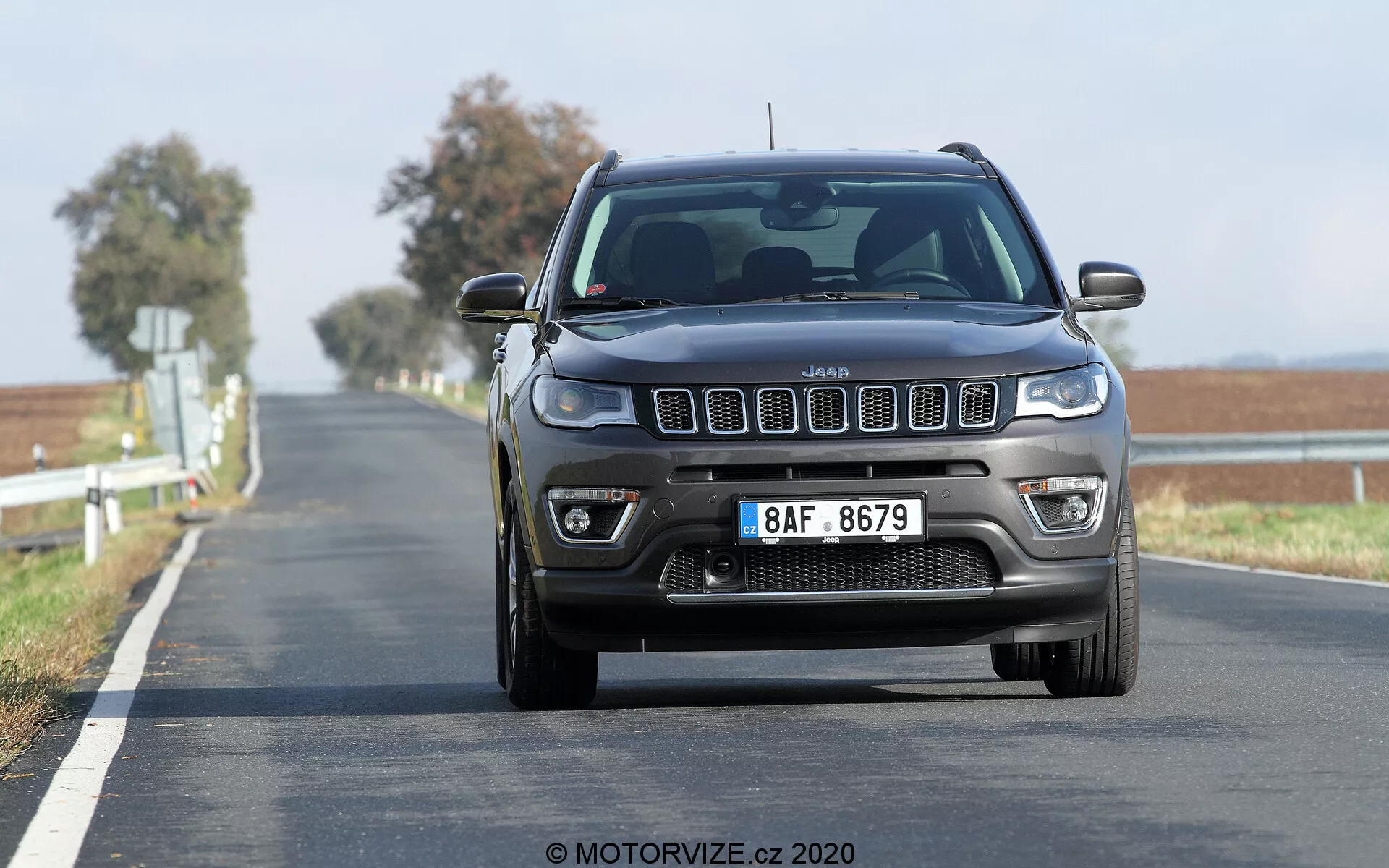 Frontalni pogled na 2017 Jeep Compass, ki se premika po dvosmerni cesti, posnet iz nizkega kota, poudarjajoč značilno sedemdelno rešetko, kvadratne žaromete z modernimi dnevnimi lučmi ter vidne meglenke in sprednje parkirne senzorje na odbijaču. Cesta se razprostira do obzorja z dvojno rumeno črto, kar označuje območje prepovedi prehitevanja. Gibajoče se zamegljeno ozadje nakazuje, da je vozilo v gibanju in ima nekoliko višji položaj, značilen za SUV-je.