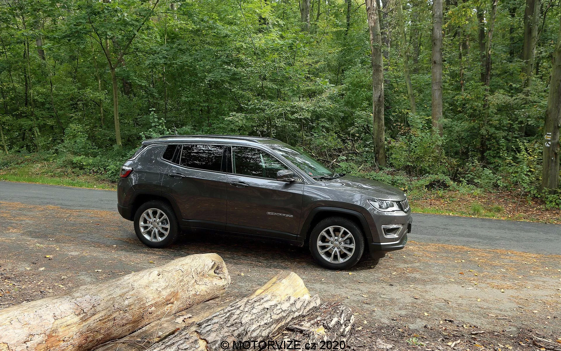 Stranski pogled na SUV Jeep Compass 2017, parkiran na asfaltni cesti v naravnem okolju z bujnimi zelenimi drevesi. Vozilo prikazuje moderne oblikovalske linije vzdolž vrat in kolesnih lokov, z vidnimi aluminijastimi platišči z večkrakim dizajnom. Ima visoko oddaljenost od tal, kar nakazuje terenske zmogljivosti. Slika zajame del sprednje rešetke in žarometov, kar kaže na sodoben slog. Prisotni so strešni nosilci, integrirani smerniki v stranskih ogledalih in zatemnjena okna. Temna barva z bleščečim zaključkom nakazuje dobro vzdrževano vozilo. Stacionarn SUV je prikazan v obilni naravni svetlobi, idealno za prikaz njegovih značilnosti in privlačnosti za ljubitelje zunanjih in avanturističnih aktivnosti.