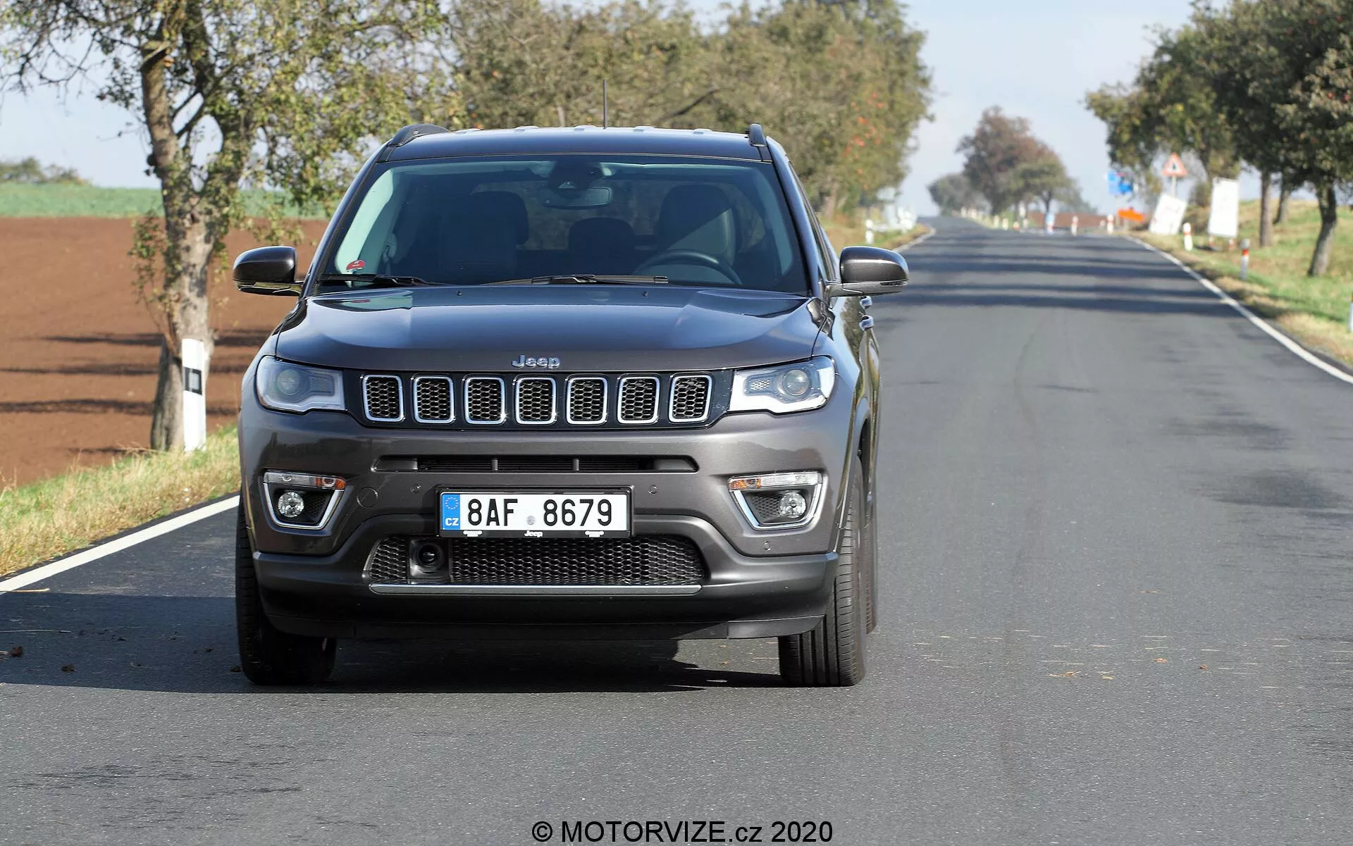 Sprednji pogled na Jeep Compass 2017, ki potuje po dvopasovni podeželski ali primestni cesti, posnet iz nizkega kota, prikazuje ikonično rešetko s sedmimi režami, LED dnevnimi lučmi, meglenkami in robustne obrise pokrova motorja. Avto je na sredini in relativno blizu kamere, z rahlim zamikom koles in ozadja. Čisto nebo kaže na svetel, sončen dan.