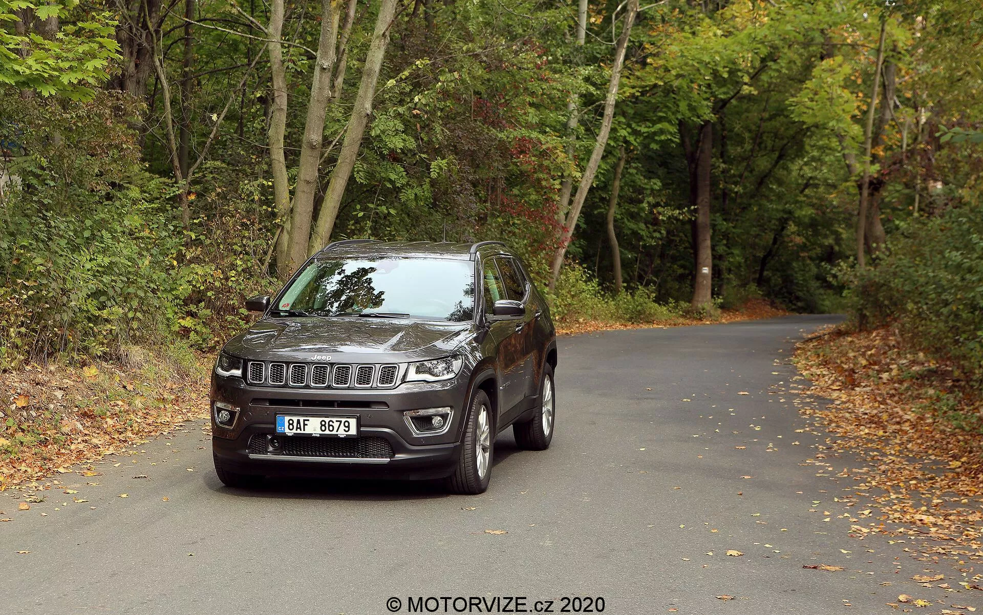 SUV Jeep Compass 2017, ki je postavljen na asfaltno cesto, obdano z gozdovi z odpadlimi listi, kar nakazuje na jesen. Vozilo je fotografirano iz sprednjega, tretjinskega kota, prikazujoc značilno Jeepovo rešetko s sedmimi odprtinami in osrednji emblem, moderne LED žaromete in meglenke, integrirane v odbijač. Opremljeno je z litimi platišči, ogledali z integriranimi smerniki, robustno izdelavo s ščitniki okoli blatnikov in rahlo zatemnjenimi stekli. Podroben pogled poudarja dizajnerske in tehnološke elemente ter nakazuje, da je vozilo primerno tako za mestne kot za terenske avanture.