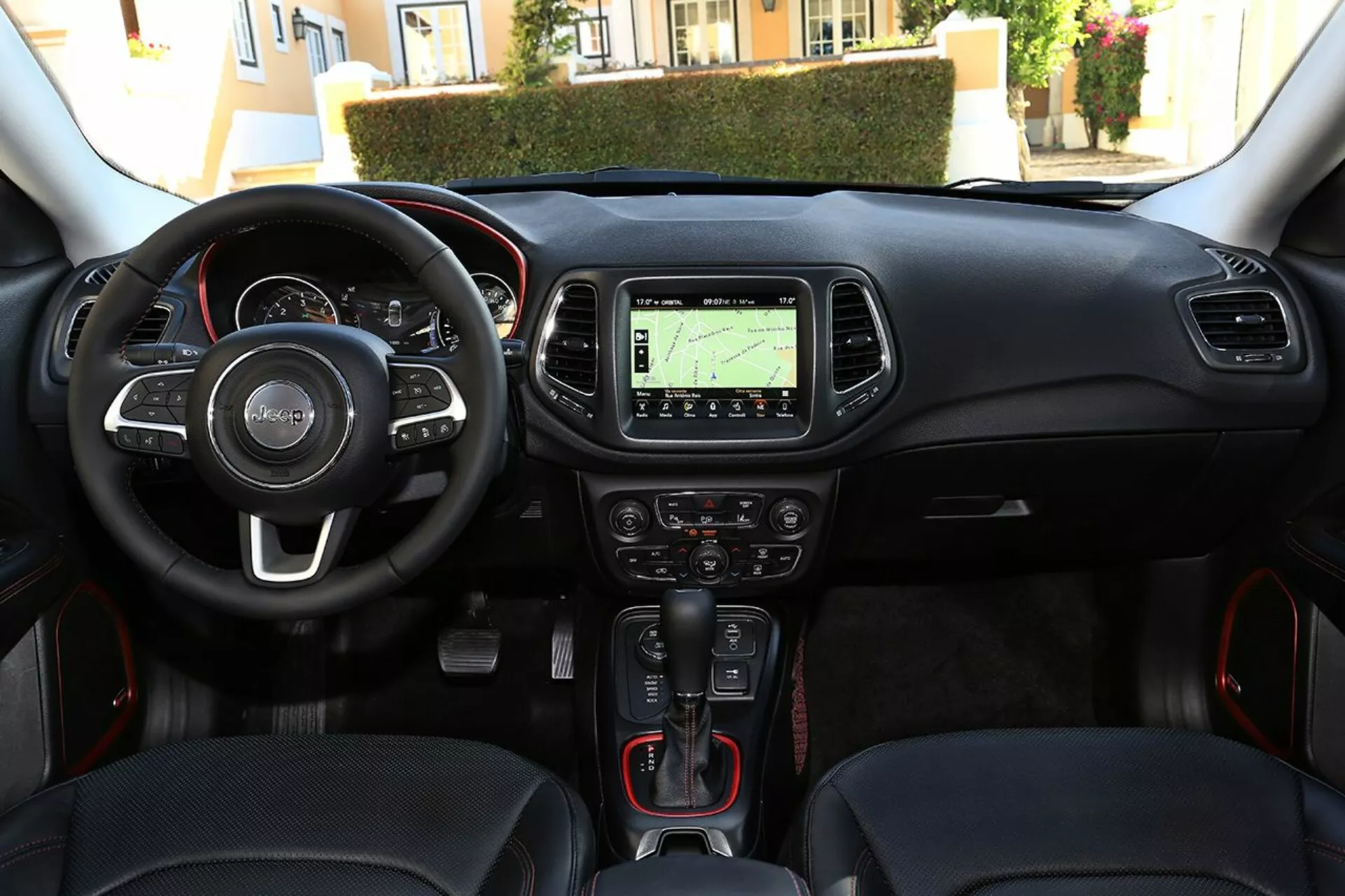 Notranji pogled na Jeep Compass 2017 iz perspektive voznika prikazuje volan z nameščenimi upravljalnimi gumbi, instrumentno ploščo, sredinsko konzolo z navigacijskim sistemom, klimatske kontrole, avtomatsko prestavno ročico z rdečimi športnimi detajli, gumb za zagon/zaustavitev brez ključa in vrata za multimedijsko povezljivost