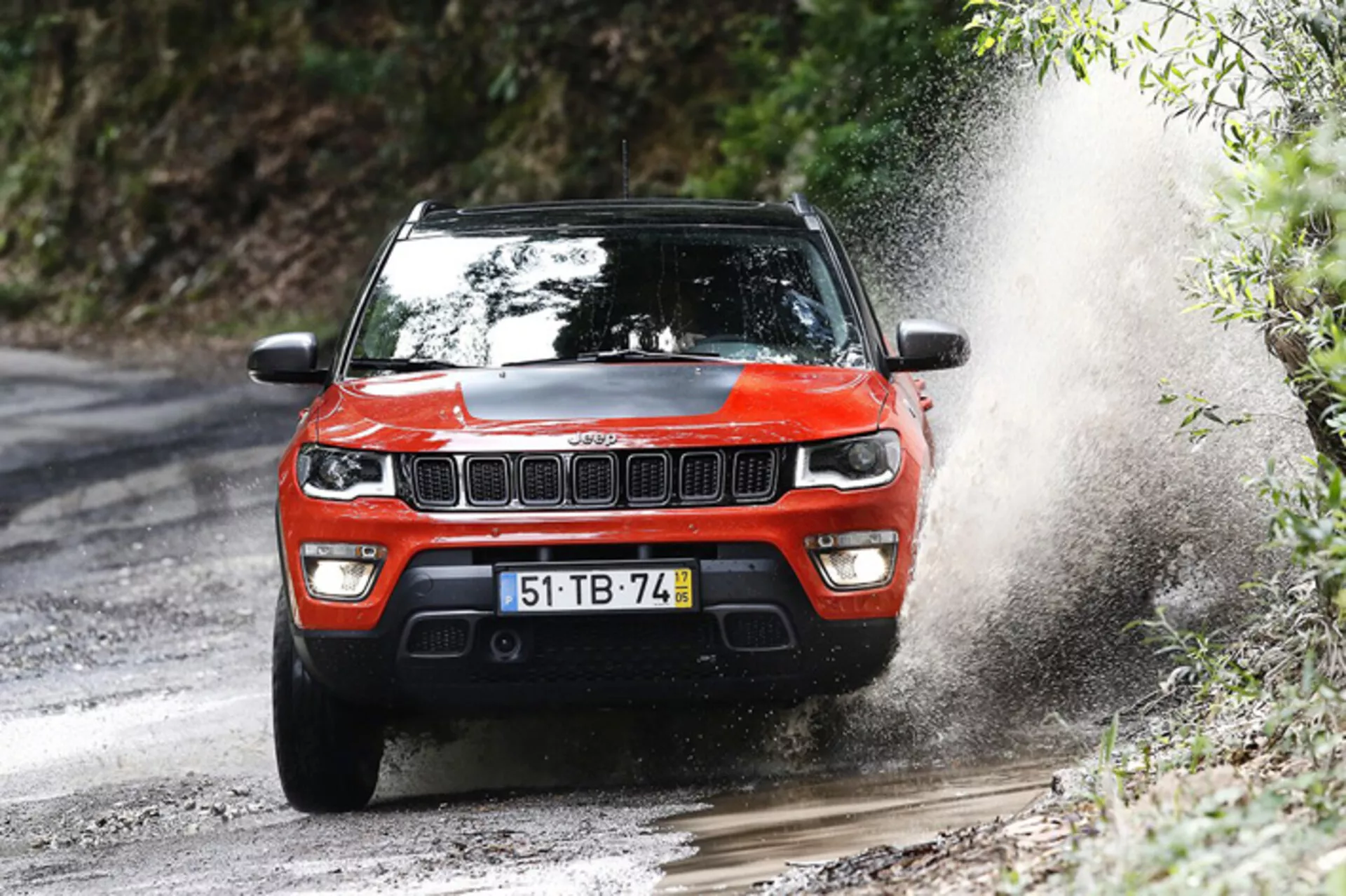 Rdeči SUV Jeep Compass letnik 2017 vozi po mokri makadamski cesti, prikazuje sprednjo rešetko, LED žaromete in terenske sposobnosti z razpršenjem vode