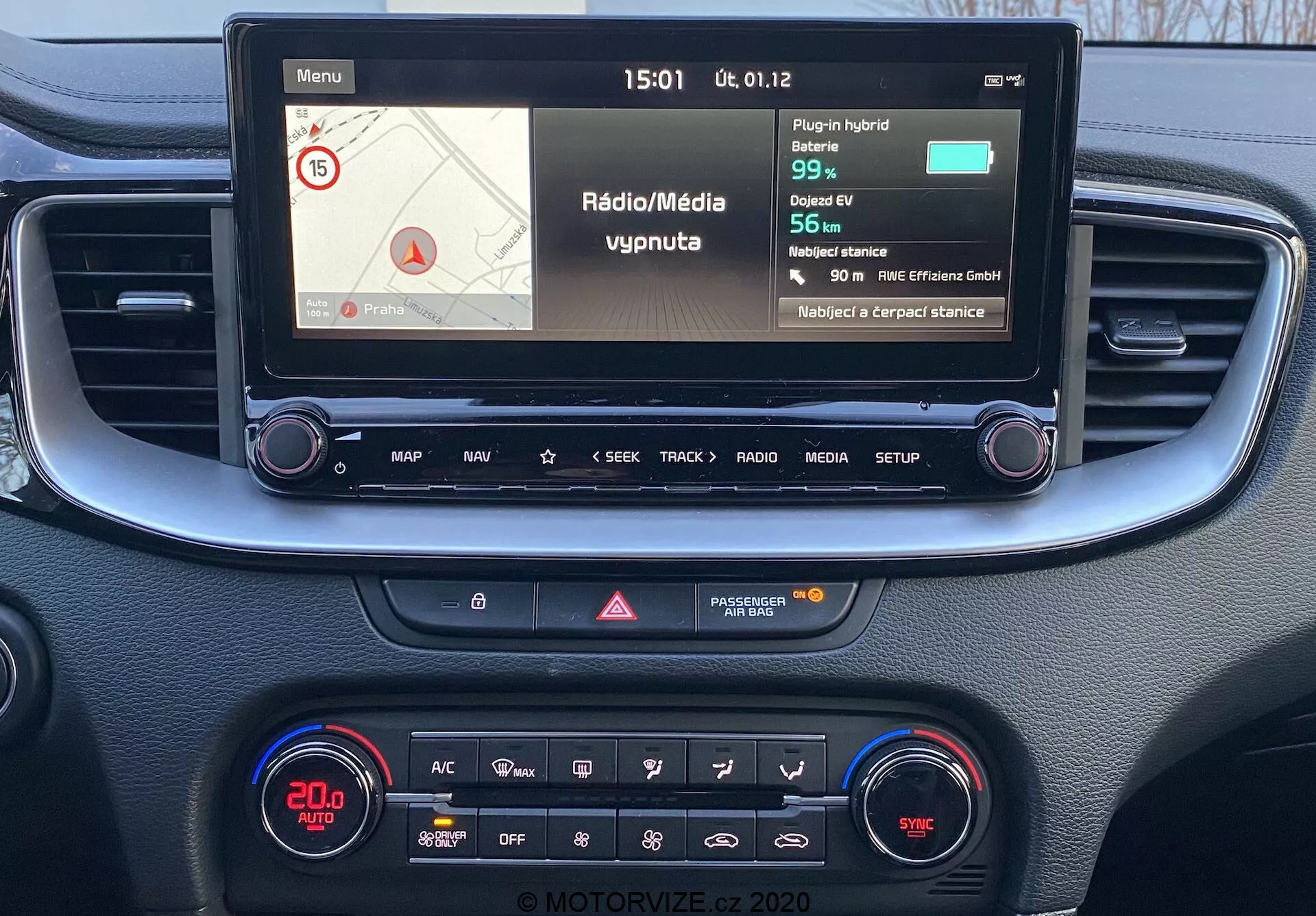 Pogled iz prve osebe na sredinsko konzolo in infotainment sistem Kia Ceed 2018 z barvnim zaslonom, ki prikazuje informacije o navigaciji in medijih, fizičnimi gumbi za nadzor in s klimatsko ploščo z digitalnim prikazovalnikom in različnimi funkcijami.