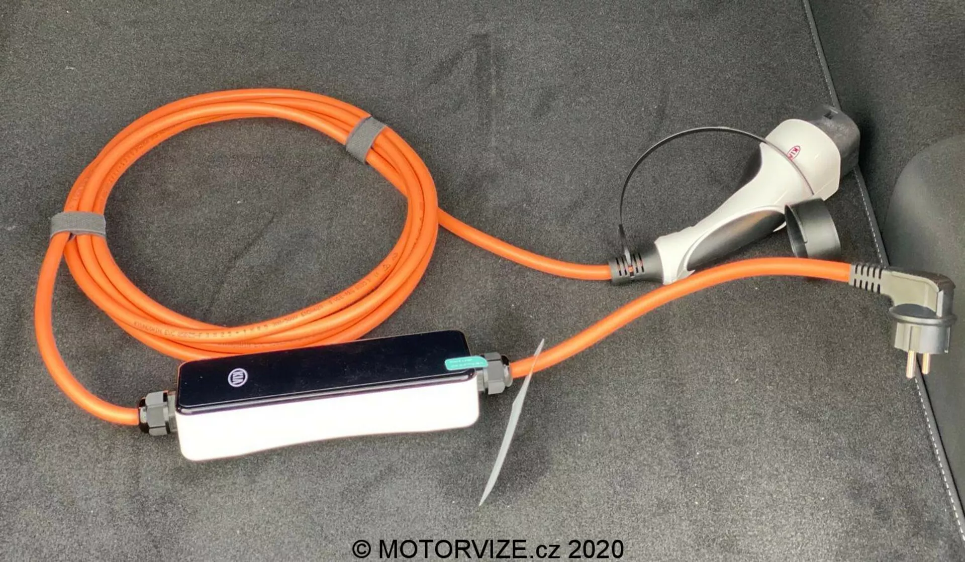 Nabojni kabel in adapter za električno vozilo (EV) Kia Ceed 2018, ki vključuje prenosljivo oranžno EVSE (Naprava za Napajanje Električnega Vozila) z visoko vidljivostjo za varnost. Konfiguracija vsebuje polnilni priključek in vtič za uporabo v domačih vtičnicah ter vmesno krmilno škatlo z LED indikatorji, vse skupaj je položeno na preprogi v prtljažniku ali tovorni površini vozila.