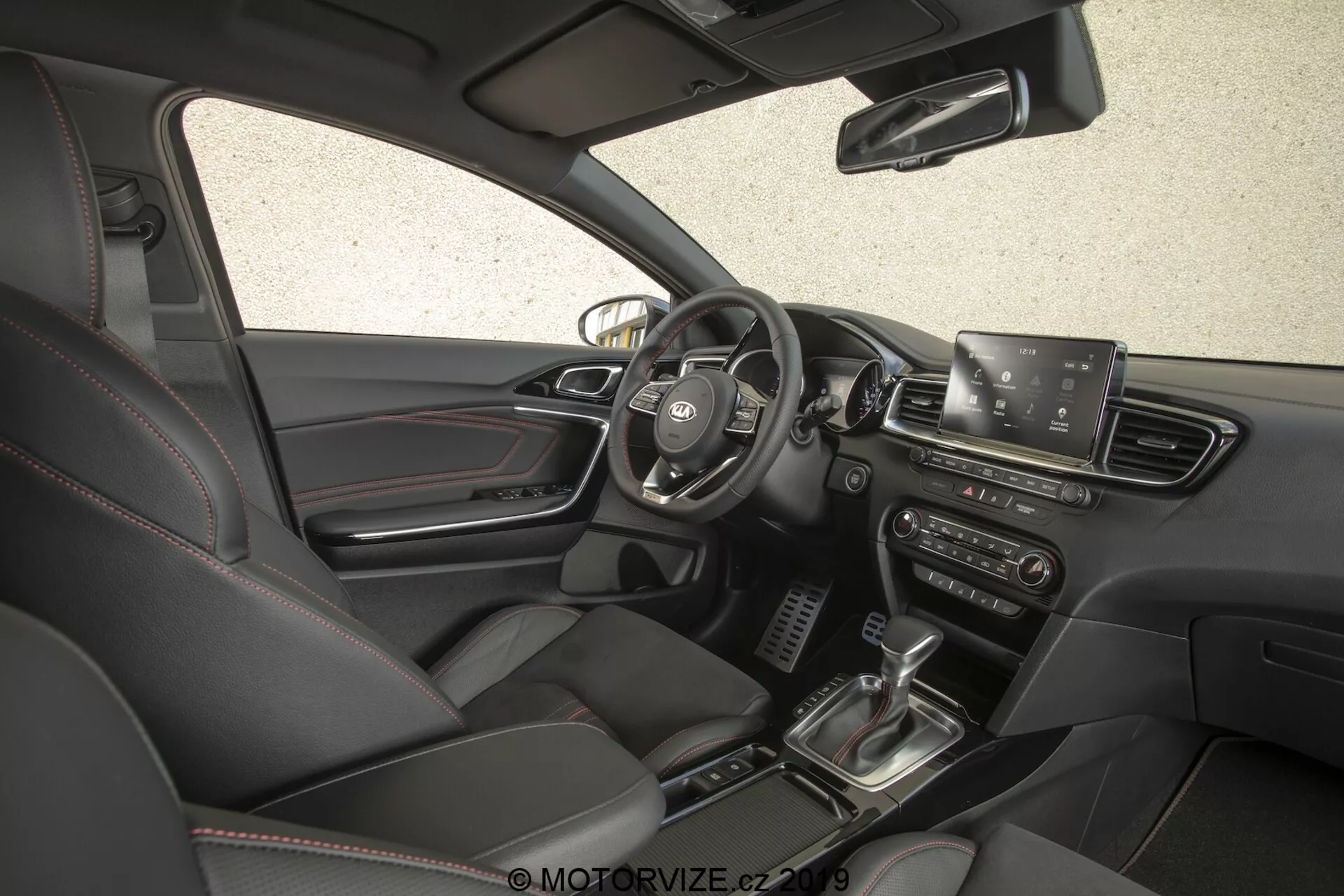 Notranji pogled avtomobila Kia Ceed 2021, ki prikazuje stran voznika iz visokega in rahlo naprej usmerjenega kota. Slika, posneta s strani potnika, zagotavlja celovit pogled na armaturno ploščo, vključno s sredinsko nameščenim volanom z nadzornimi gumbi, delno vidljivostjo instrumentne plošče, vidnim infotainment zaslonom, zračniki, gumbi in gumbi za nadzor klime, prestavno ročico, sistemom zagona na gumb, usnjenimi sedeži z električnimi nastavitvami in temno barvno paleto notranjosti.