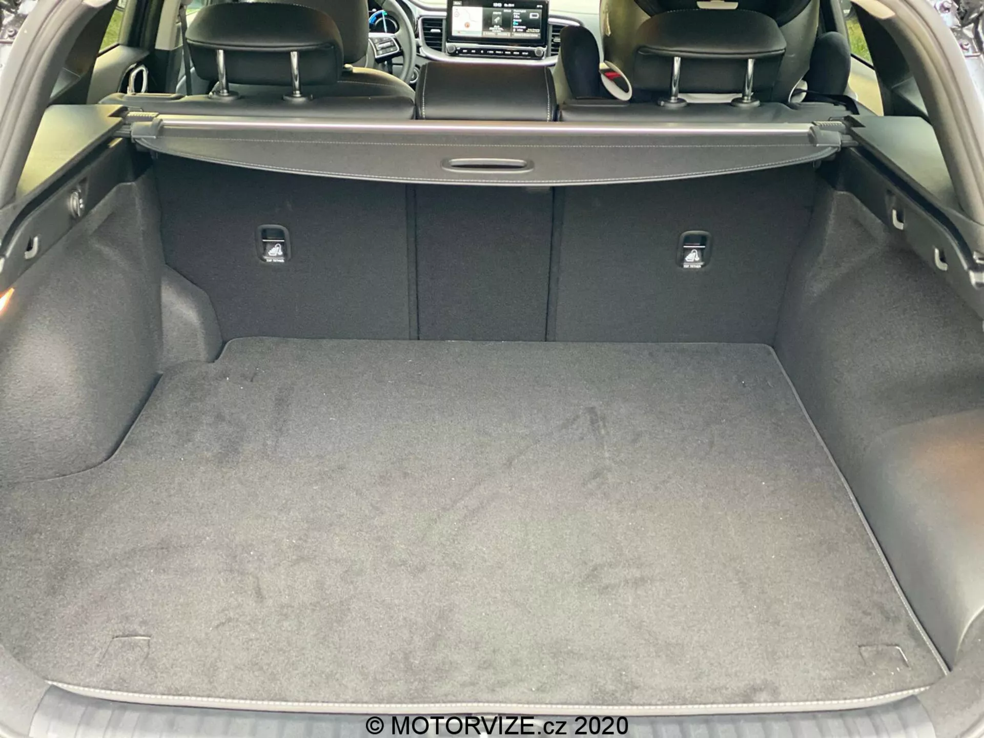 Bližnji pogled na prtljažnik Kia Ceed 2018 iz zadnje perspektive, poudarjajoč prostorno in čisto prtljažno področje s preprogo. Prtljažna vrata so dvignjena, kar razkriva izvlečno pokrivalo prtljažnika, naslonjala zadnjih sedežev temnejše barve, verjetno iz usnja, in srebrna vlečna jezička za zlaganje sedežev. Strani so obložene z mehkim materialom, na desni strani je viden majhen predal. Slika je posneta iz rahlega kota, prikazuje dizajn in materiale notranjosti prtljažnika avtomobila.