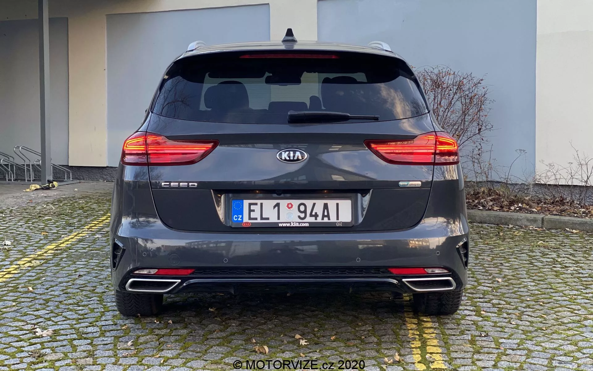 Pogled od zadaj na Kia Ceed 2018 parkirano na tlakovani površini blizu pločnika. Avto, fotografiran podnevi iz ravnega zornega kota na višini oči, prikazuje razdeljene LED zadnje luči, zadnje steklo z brisalcem, oznako proizvajalca, ime modela, registrsko tablico, zadnji odbijač z odsevniki in parkirnimi senzorji, dvojni izpušni sistem in kovinsko temno sivo barvo. V ozadju je stavba, kar nakazuje na stanovanjsko ali komercialno območje.