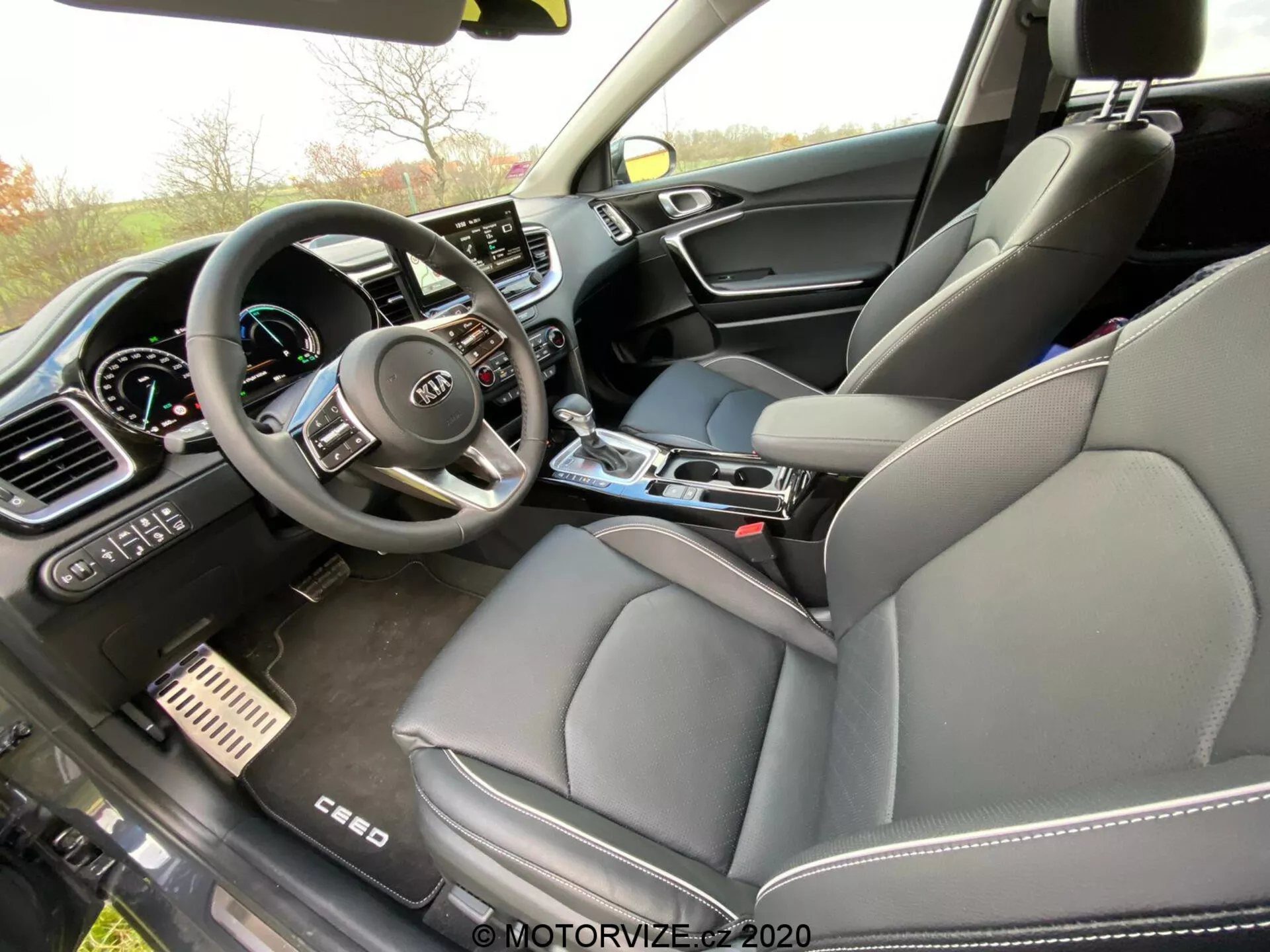 Posnetek notranjosti Kia Ceed 2018 iz potniške perspektive pod kotom, ki zajema sprednji del s poudarkom na armaturni plošči, sredinski konzoli in sedežu voznika. Volan z več kontrolnimi gumbi in logotipom Kia je jasno viden, skupaj z delno videnim digitalnim instrumentnim zaslonom. Sredina armaturne plošče vsebuje velik zaslon na dotik informacijsko-razvedrilnega sistema, obdan z ročnimi nastavitvami klimatizacije. Sredinska konzola vključuje prestavno ročico, držala za pijačo in kontrolne gumbe. Vidna sta tudi sprednji sedež potnika s usnjeno oblazinjenjem in mešanimi materiali na vratnih panelih, kar nakazuje na luksuzno opremo. Kovinski pedal za zavoro in pospeševanje prav tako nakazujeta premium športni pridih.