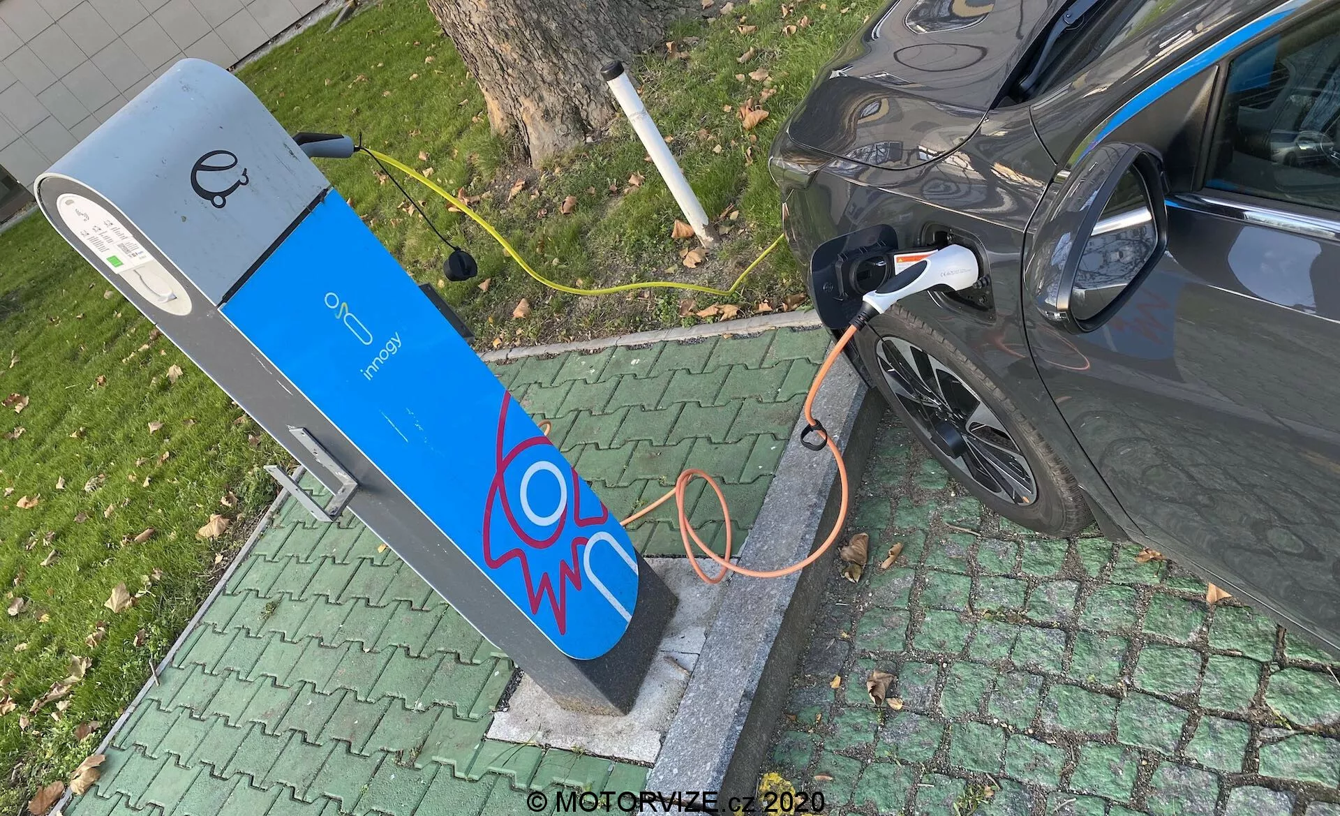 Visok kotni pogled na sodoben električni avtomobil, ki se polni na javni polnilni postaji. Slika prikazuje desni zadnji del avtomobila, osredotočen na polnilni priključek s priključenim debelim oranžnim kablom. Polnilna postaja ima modro fasado z motivom električne iskrice in zaslonom na vrhu, ki nakazuje elemente uporabniškega vmesnika. Prizor je postavljen v zeleno, parkovno okolje, kar nakazuje priročno urbano ali primestno lokacijo.