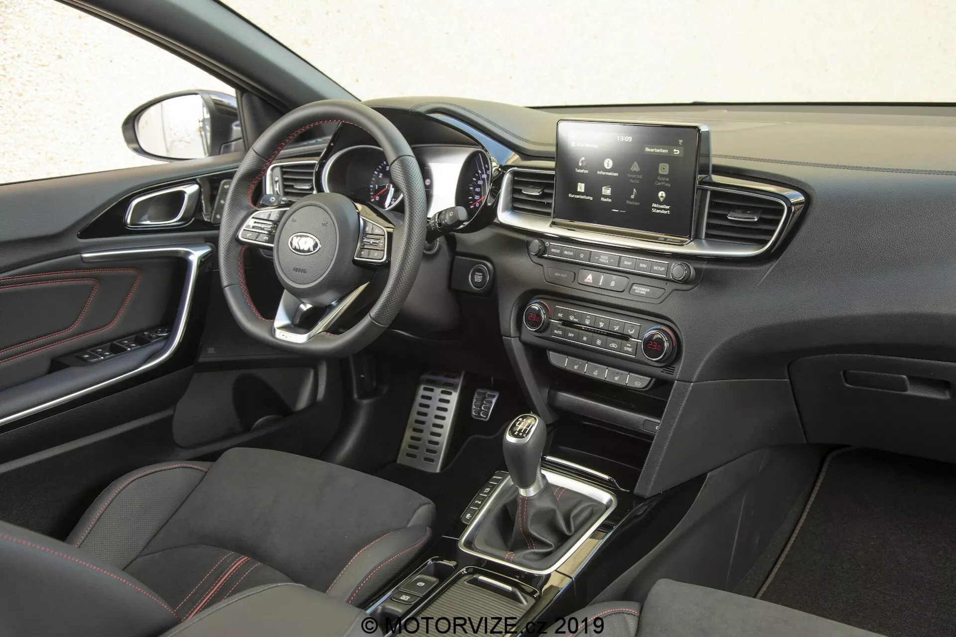 Notranjost Kia Ceed 2021, osredotočena na voznikovo stran iz bočne perspektive. Volan z prepoznavnim logotipom proizvajalca ima večfunkcijske kontrole za upravljanje infotainmenta, tempomata, telefonske povezljivosti in morda glasovnimi ukazi. Za volanom se nahajajo merilnik hitrosti, tahometer in merilniki informacij za voznika. Sredinska konzola ima zaslon na dotik za infotainment, ki ponuja navigacijo, nastavitve vozila, predvajanje medijev in povezljivost Bluetooth. Spodaj so ročne kontrole za avdio in klimatske sisteme. Ročica ročnega menjalnika, obdana z gumbi za načine vožnje ali nadzor zdrsa, in športni sedeži z usnjenimi in tkaninskimi vložki povečujejo občutek zmogljivosti in prestiža. Kovinske stopalke v voznikovem podnožju kažejo na poudarek na zmogljivosti, medtem ko kromirani detajli na odprtinah za nadzor klime, kljukah za vrata in drugih podrobnostih dodajajo prefinjenost. Poklopec za vrata vključuje kontrole za električna okna in morda nastavitve električnih ogledal ter je vidni del vzvratnega ogledala, kar nakazuje funkcije, kot so samodejno zatemnjevanje ali vgrajen kompas.