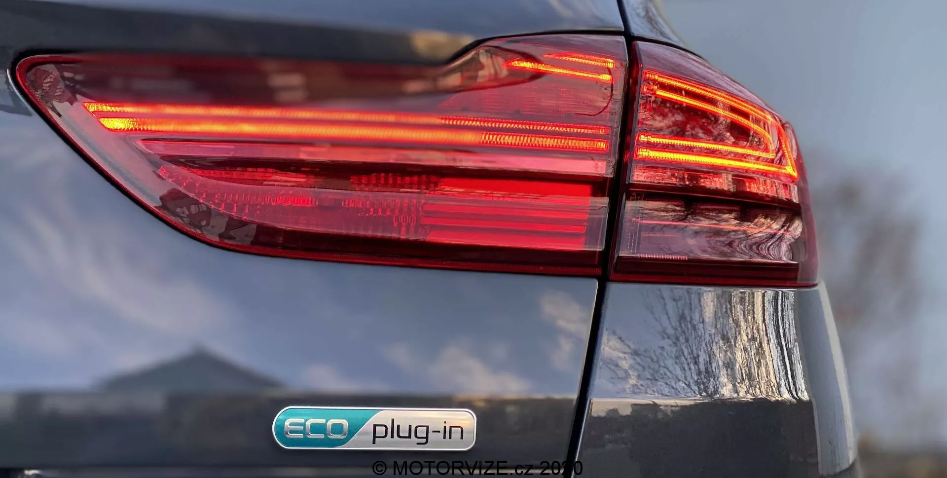 Od blizu prikazan zadnji levi del Kia Ceed 2018, s poudarkom na osvetljenem zadnjem svetlobnem sklopu z večplastno zasnovo. Značka 'ECO plug-in' označuje, da gre za priključno hibridno vozilo. Sijajni zaključek in delno viden pokrov prtljažnika dodajata podrobnosti celotni zasnovi avtomobila.