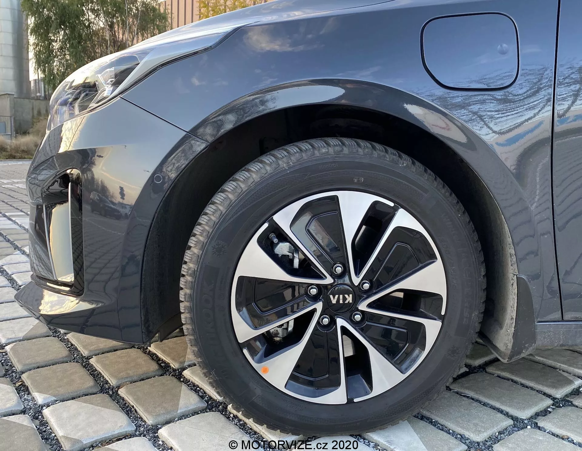 Pogled od blizu iz nizkega kota na desno prednje kolo KIA Ceed 2018, z večkrakim aluminijastim dizajnom v črni in srebrni barvi, z vidnim vzorcem tekalne plasti, ki delno prikazuje prednji blatnik, odbijač in del pokrova motorja v temni kovinski barvi.