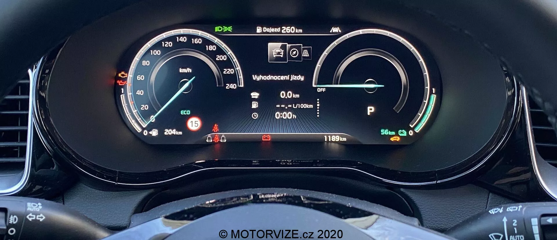 Pogled od blizu na digitalno armaturno ploščo Kia Ceed 2018 iz voznikovega sedeža. Zaslon vključuje merilnik hitrosti s fizično iglo in digitalnim prikazom, ki kaže 15 km/h. Na desni strani je merilnik moči/baterije, ki kaže popolnoma napolnjeno baterijo z dosegom 481 km. Drugi elementi vključujejo zaslon pripravljenosti, informacije za voznika z opozorilnimi lučkami, zunanjo temperaturo 20,5°C in vozilo v načinu Park. Vidni so tudi zgornji lok volana s športnim, ploskim spodnjim delom.