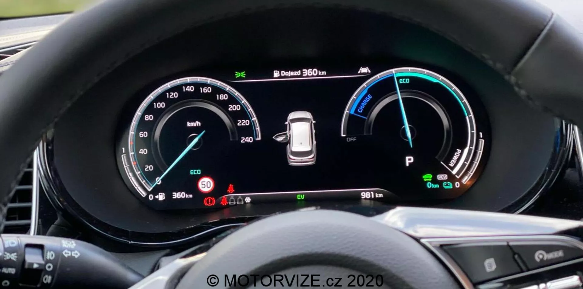 Pogled od blizu na digitalno instrumentno ploščo Kia Ceed 2018 z voznikove perspektive, z velikim širokozaslonskim prikazom in podrobnimi instrumenti, vključno s hitrostomerom do 260 km/h, merilnikom moči z načini Eco, Comfort in Power, opozorilom o tlaku v pnevmatikah in indikatorjem načina 'Park'. Instrumentna plošča prikazuje tudi 360-stopinjski kamerni sistem in števec kilometrov z odčitkom 9811 km. Obloga zmanjša bleščanje, gumbi na volanu so delno vidni. Vodni žig 'MOTORVIZE.cz 2020'.