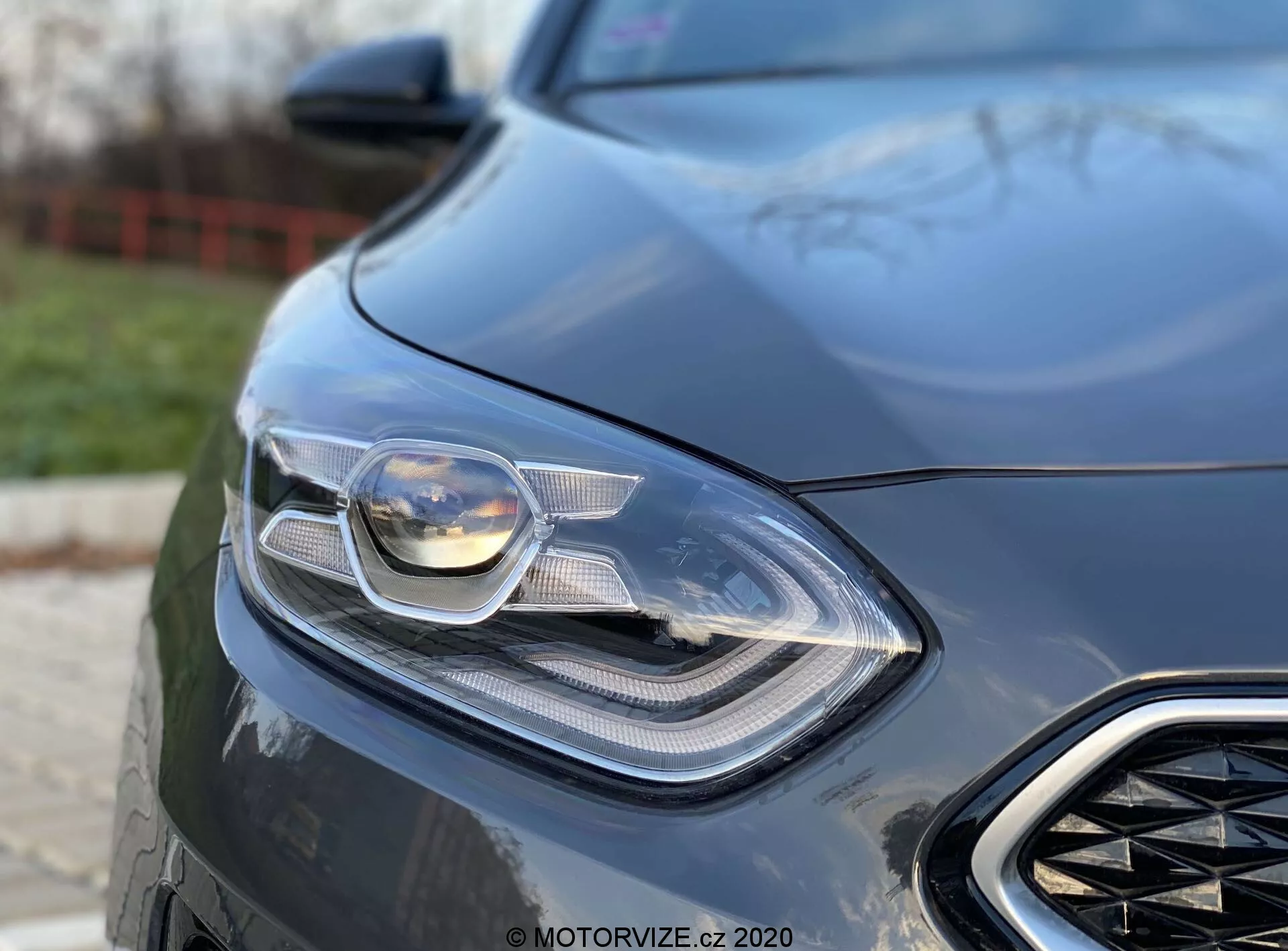 Podroben bližnji posnetek levega sprednjega dela Kia Ceed 2018, osredotočen na sofisticiran žaromet in del sprednje mreže. Posnetek je narejen iz nizkega kota, kar poudarja eleganten in moden dizajn avtomobila z vidno napredno osvetljevalno tehnologijo, ki vključuje LED in prilagodljive žaromete. Zamegljeno ozadje poudarja aerodinamične elemente avtomobila in občutek razkošja, kar nakazuje na zmogljivostno naravnano vozilo.