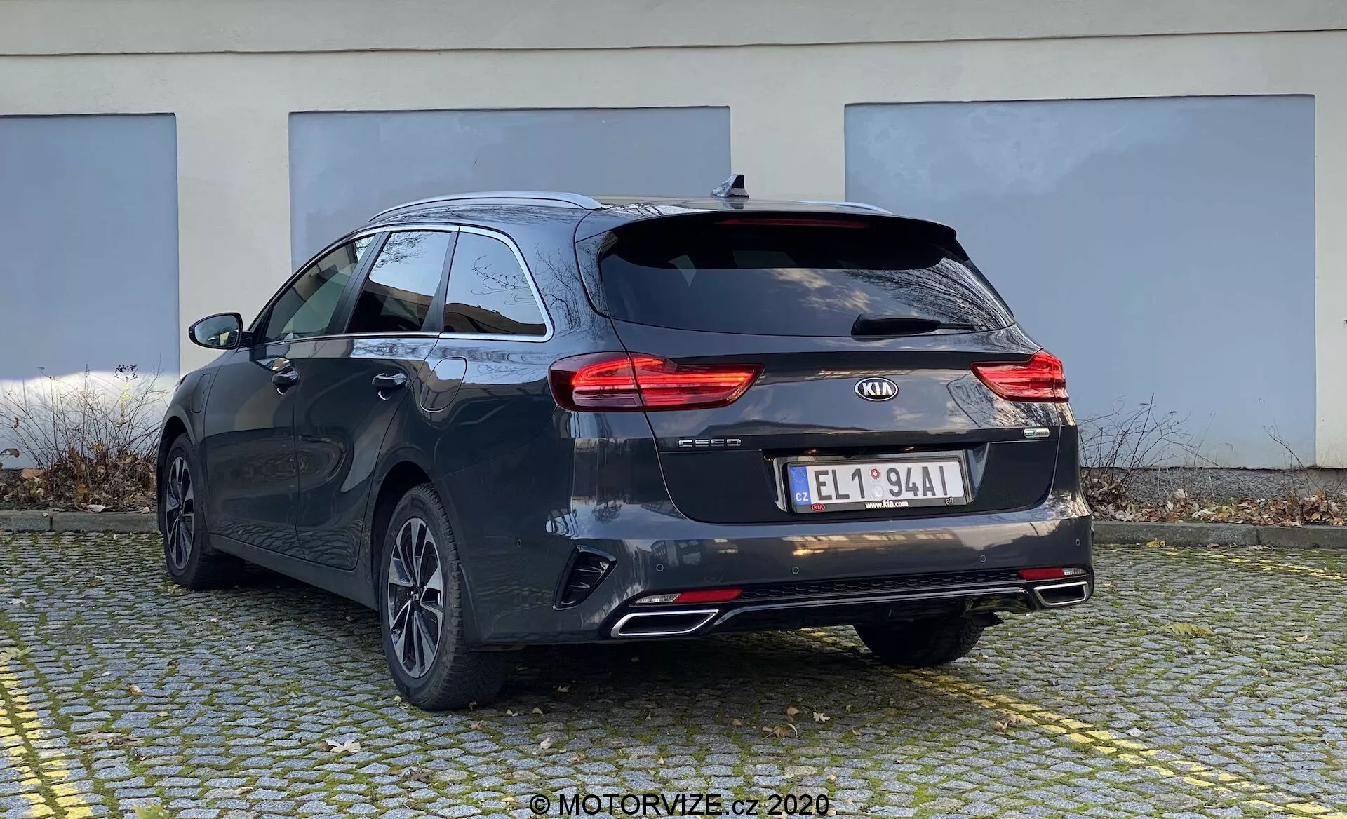 Slika prikazuje Kia Ceed 2018 iz zadnje tričetrtinske perspektive, pri čemer hkrati poudarja zadnji del in stran avtomobila. Vozilo je v temni barvi, verjetno črni ali temno sivi, z značilnimi linijami in rahlo padajočo streho za športni videz. Vidne so moderne LED zadnje luči, logotip Kia, ime modela 'CEED', dodatne oznake, evropska registrska tablica, zadnja kolesa večkraka aluminijastih platišč, dvojne izpušne cevi in zadnji brisalnik. Predvideva se, da so vgrajena vzvratna kamera in parkirni senzorji, vendar niso vidni. Majhen strešni spojler in strešni nosilci kažejo na dodatne možnosti za prtljago. Avto je parkiran na tlakovanem območju z zgradbo v ozadju, prikazuje zadnji del in del bočnega profila iz rahlo nizkega kota.