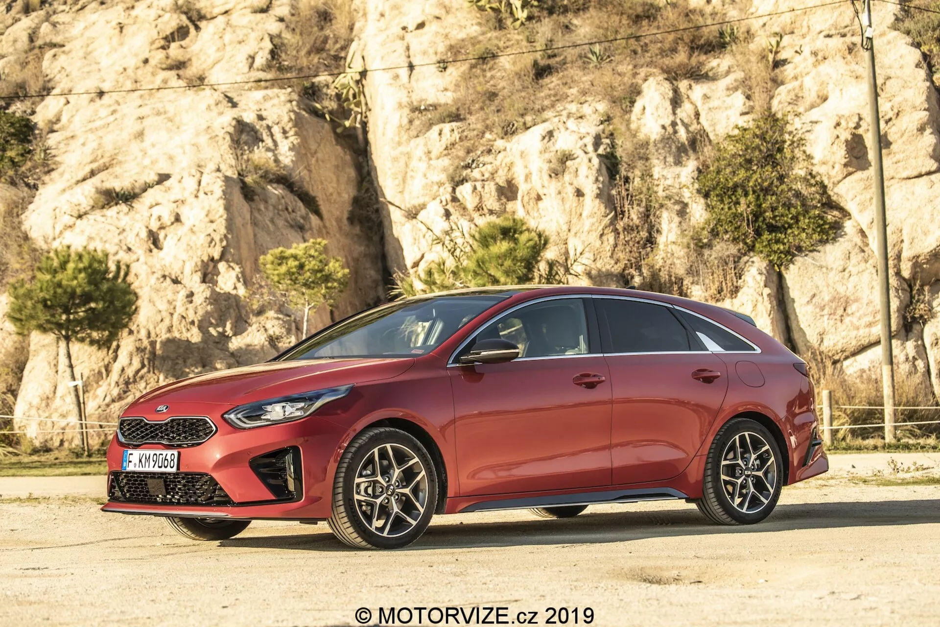 Stranski profil rdeče hatchback Kia Ceed 2021, z elegantno, aerodinamično obliko z nagnjenimi linijami strehe in oblikovanimi linijami na vratih. Avto, petvratni model, ima kromirano obrobo okna in zatemnjene stebričke B, kar ustvarja učinek neprekinjene steklene plošče. Večkraka platišča iz lahke litine, z vidnimi kolutnimi zavorami, dodajajo športen pridih. Ostre žaromete in delno vidna mreža označujejo sprednji del, medtem ko ogledala v barvi karoserije z vgrajenimi smerniki izboljšujejo vidljivost in varnost.