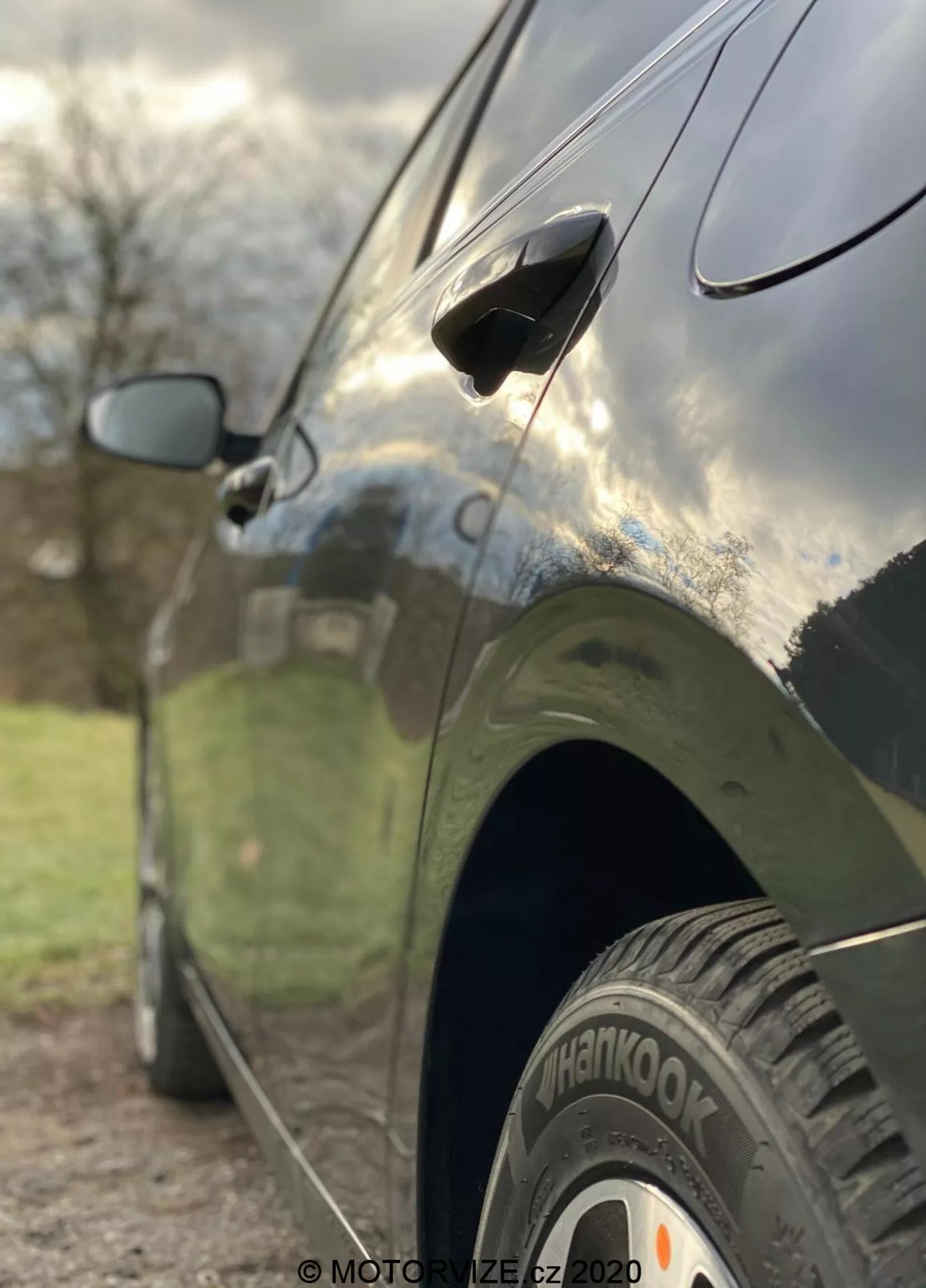 Od bliže je prikazan stranski pogled na Kia Ceed 2018, poudarjajoč značilnosti, kot so zadnje kolo s pnevmatiko 'Hankook', aerodinamičen ročaj na vratih in delno zamegljeno stransko ogledalo. Fotografija je posneta iz nizkega kota z majhno globinsko ostrino, ki ostro izostri zadnje kolo in spodnji del vrat, medtem ko je ozadje umetniško zamegljeno. Avto ima temen, sijoč zaključek, ki odraža nebo in okolico, s čimer poudarja elegantne oblikovne linije in dobro vzdrževano karoserijo.