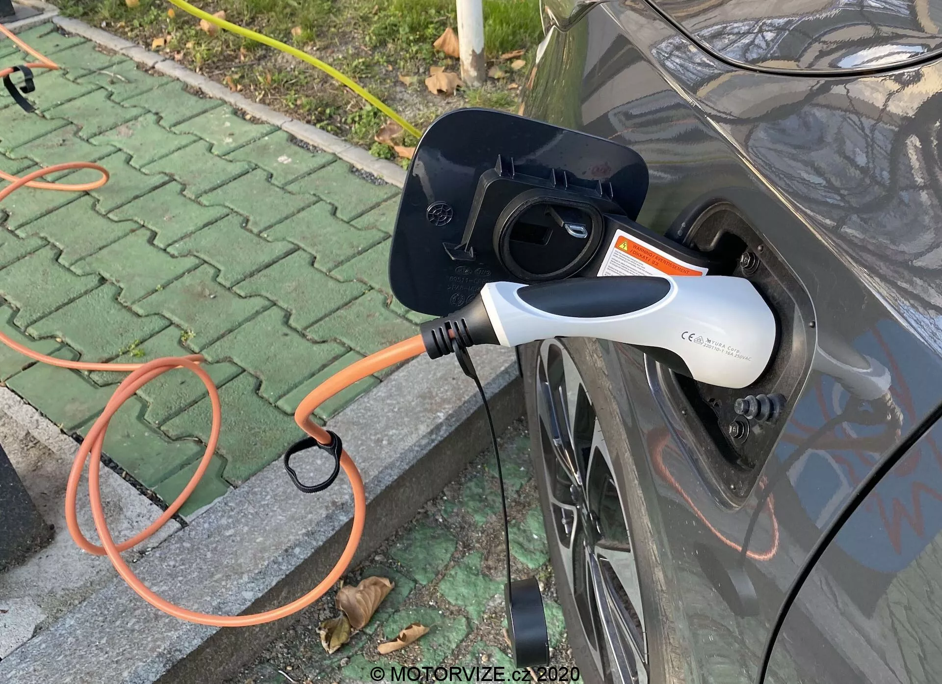 Bližnji stranski pogled na sivo električno vozilo (EV) med polnjenjem, z poudarkom na odprtem polnilnem vratu in priključenem oranžnem EVSE priključku. Sodobni beli in črni polnilni priključek ima LED indikator. Sliko se osredotoča na interakcijo med avtomobilom in polnilno infrastrukturo, umeščeno v zunanji tlakovan prostor z raztresenimi listi.