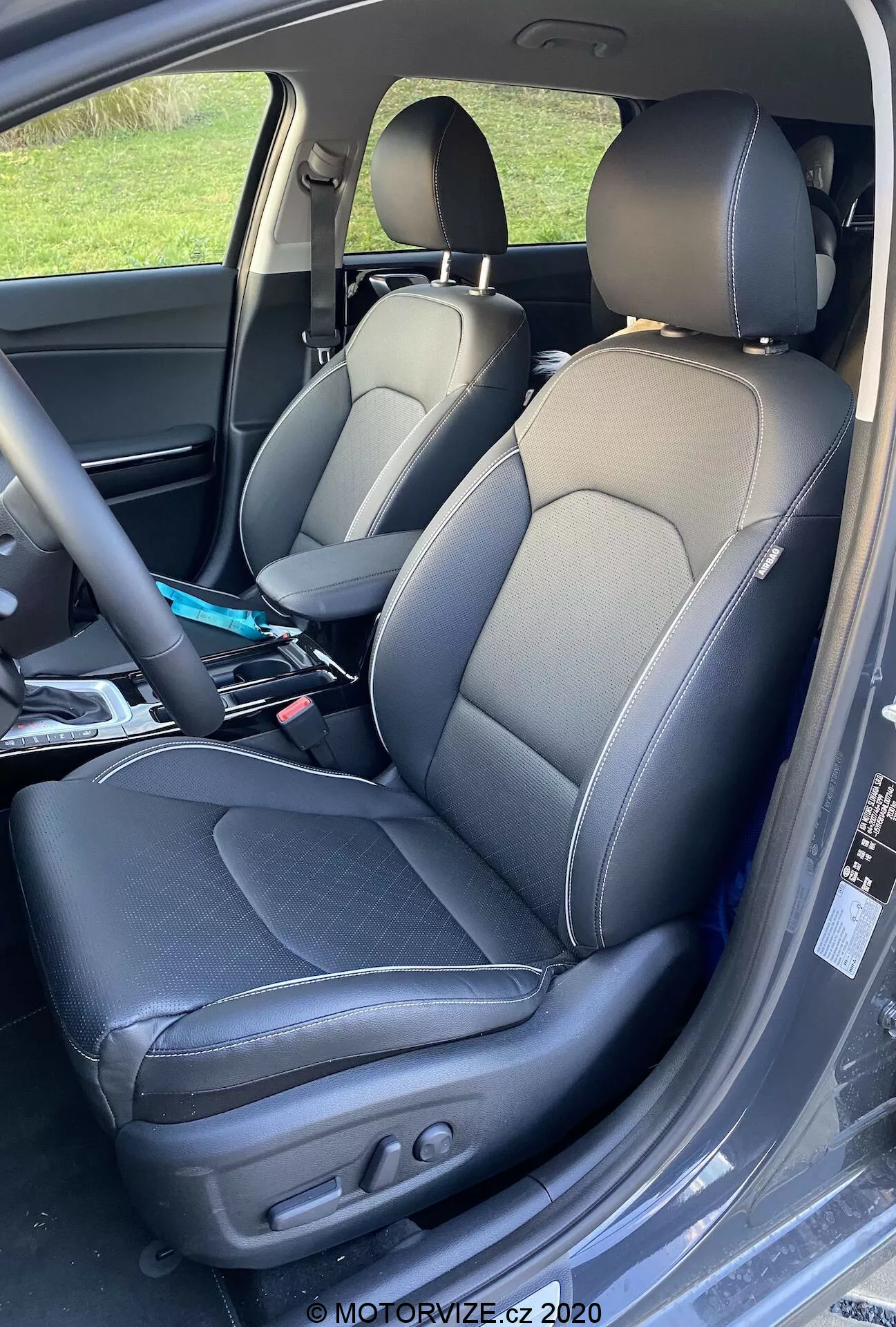 Bližnji posnetek notranjosti Kia Ceed 2018, ki prikazuje voznikov in sovoznikov sedež z dvobarvnim usnjenim oblazinjenjem s kontrastnimi šivi, športni dizajn sedežev z bočnimi oporami in električne nastavitve na voznikovem sedežu za nadzor naklona naslona in položaja sedeža. Lažni gubi kažejo na uporabo, na sovoznikovem sedežu pa so vidni različni predmeti.