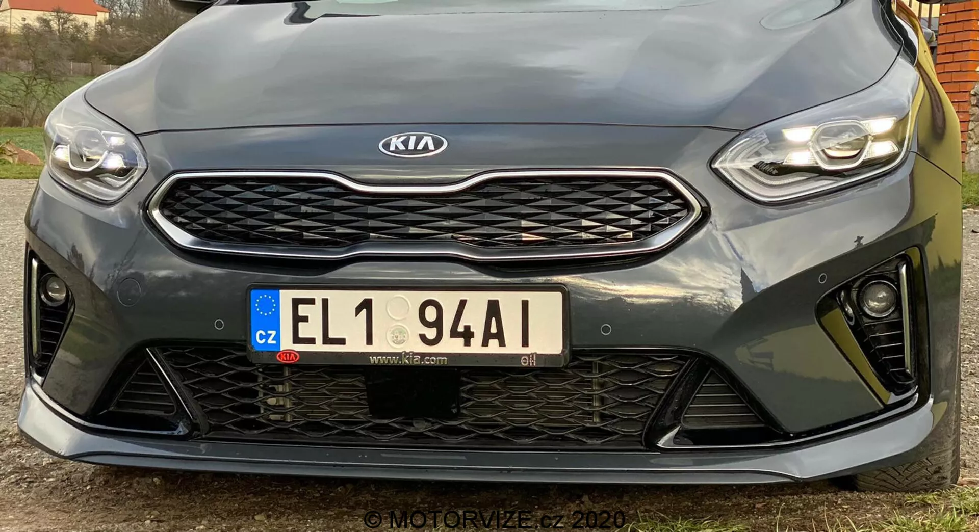 Ta slika prikazuje sprednji del vozila Kia Ceed 2018, posnetega iz nizkega kota. Poudarja elegantne, vodoravno orientirane LED žaromete, izrazito mrežo z vzorcem satja z osrednjim Kia emblemom, evropsko registrsko tablico iz Češke republike in športni sprednji odbijač z velikimi odprtinami. Bližnji pogled poudarja parkirne senzorje pod mrežo. Športni dizajn nakazuje višjo ravnino opreme, čeprav natančne specifikacije niso jasne. Visokoločljivostna slika zajema oblikovne podrobnosti in konture, s rahlo premaknjeno kotom v levo.