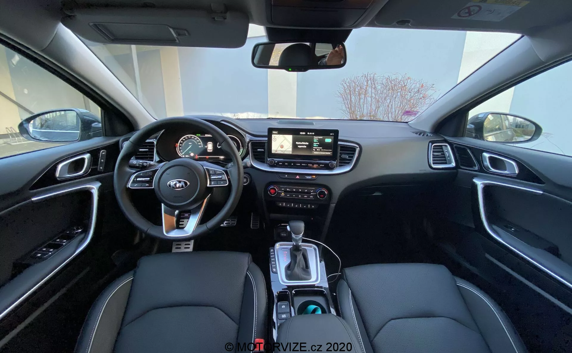 Širokokotni pogled na notranjost Kia Ceed 2018 s sedeža zadaj, ki prikazuje voznikov del, vključno z volanom z nameščenimi kontrolami, instrumentno ploščo z digitalnimi in analognimi merilniki, osrednjim info-zabavnim sistemom z zaslonom na dotik, ročnimi kontrolami za info-zabavno napravo in klimo ter osrednja konzola z prestavno ročico, izbiro načina vožnje, držali za pijače in notranjost v črni barvi s sedeži iz sintetičnega usnja z belimi šivi in oblogo iz brušenega aluminija.