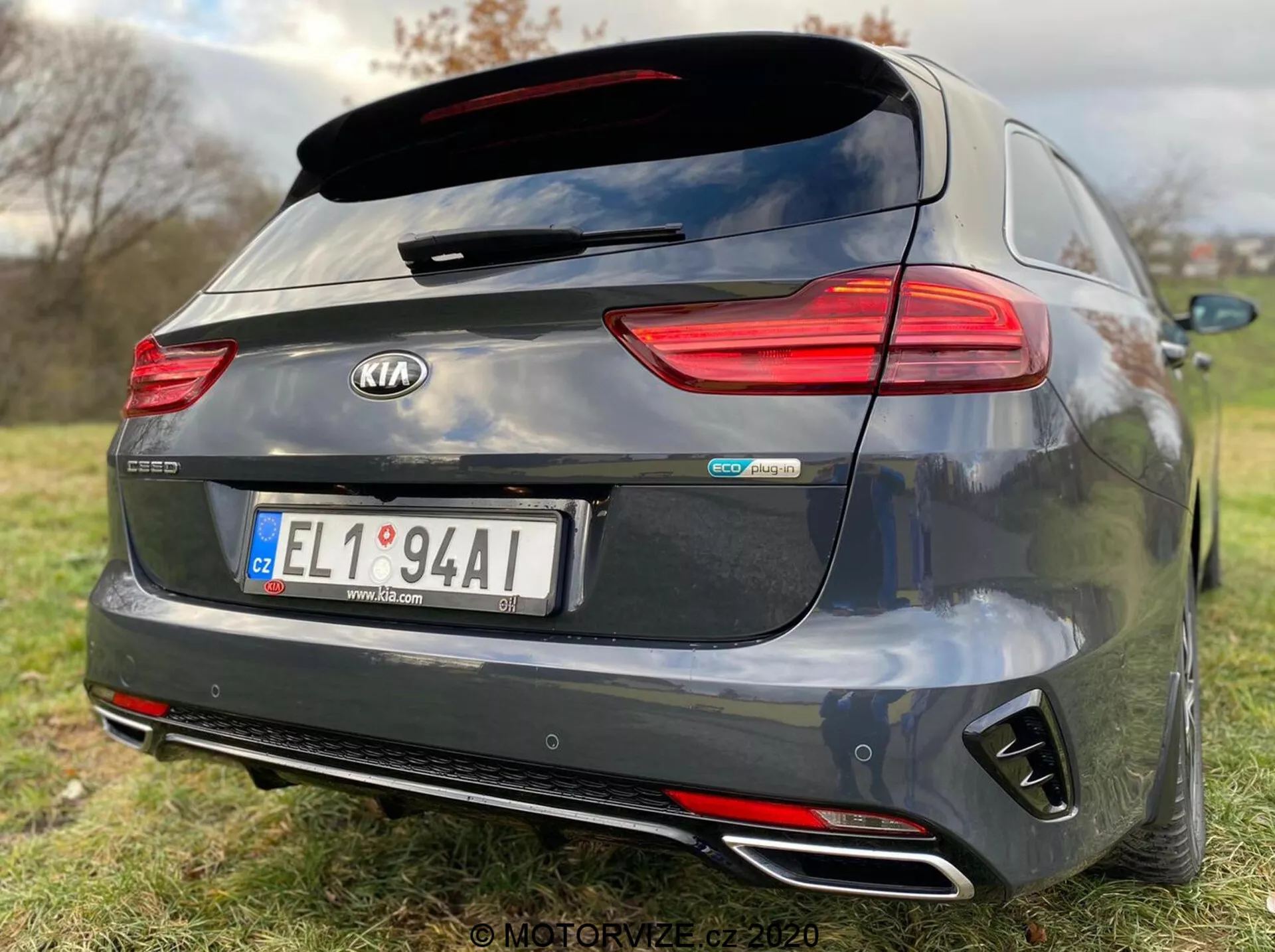 Zadnji pogled na temno siv model Kia Ceed Proceed 2018, z detalji, kot so vodoravne LED zadnje luči, strešni zadnji spojler, dvojne kromirane izpušne cevi, zadnji odbijač v stilu difuzorja in osrednji znački 'Kia' in 'GT Line'. Zadnja kamera nad osrednje nameščeno registrsko tablico nakazuje pomoč pri parkiranju, plitka globinska ostrina pa poudarja znamko in identifikatorje modela avtomobila na travnatem ozadju.