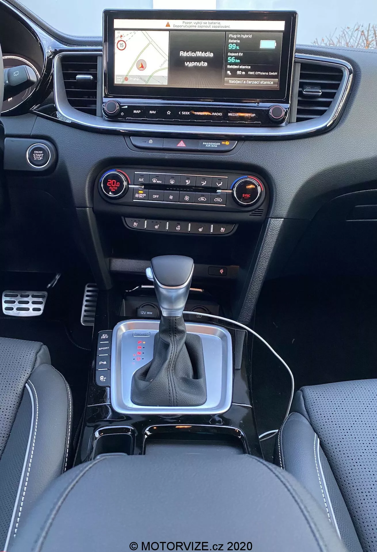 Notranji pogled Kia Ceed 2018 iz perspektive zadnjega sedeža, prikazuje moderen armaturni sistem s zaslonom na dotik, prikazuje navigacijski zemljevid, fizične gumbe in gumbi za uravnavanje avdio in klimatskega sistema, centralno prezračevalno rešetko, prestavno ročico v nevtralnem položaju s srebrnim poudarkom in črno usnjeno boot, majhen prostor za shranjevanje blizu prestavne ročice, verjetno za 12V vtičnico ali USB priključke, usnjeni sedeži z natančnimi šivi.