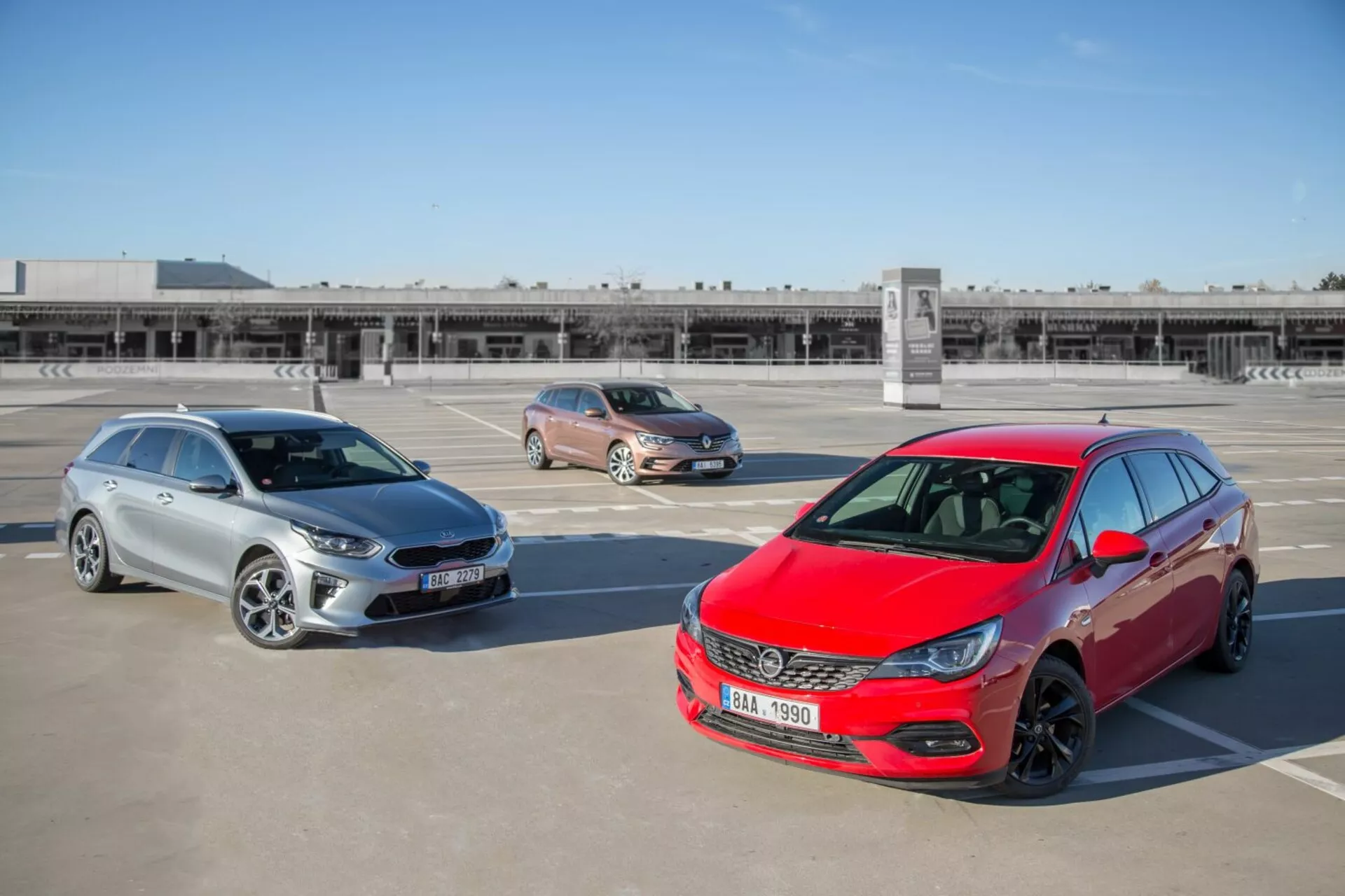 Trije modeli Kia Ceed iz leta 2009, vključno z rdečim hatchbackom, zlatim križancem in srebrnim karavanom, parkirani na strešnem parkirišču, fotografirani iz dvignjene pozicije, ki prikazuje sprednji dizajn, stranske profile in dolžino vozil