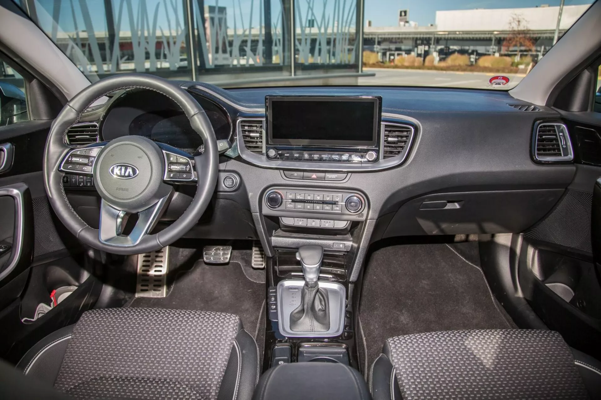 Notranjost Kia Cee'da iz leta 2009, ki prikazuje celotno sprednjo kabino, vključno z večfunkcijskim volanom z logotipom Kia, delom instrumentne plošče za volanom, prestavno ročico v parkirnem položaju na sredinski konzoli, gumbi za nadzor klime, zaslonom za infotainment, sedeži s tkaninsko oblogo in kontrolami vrat voznika in sopotnika v naravni dnevni svetlobi