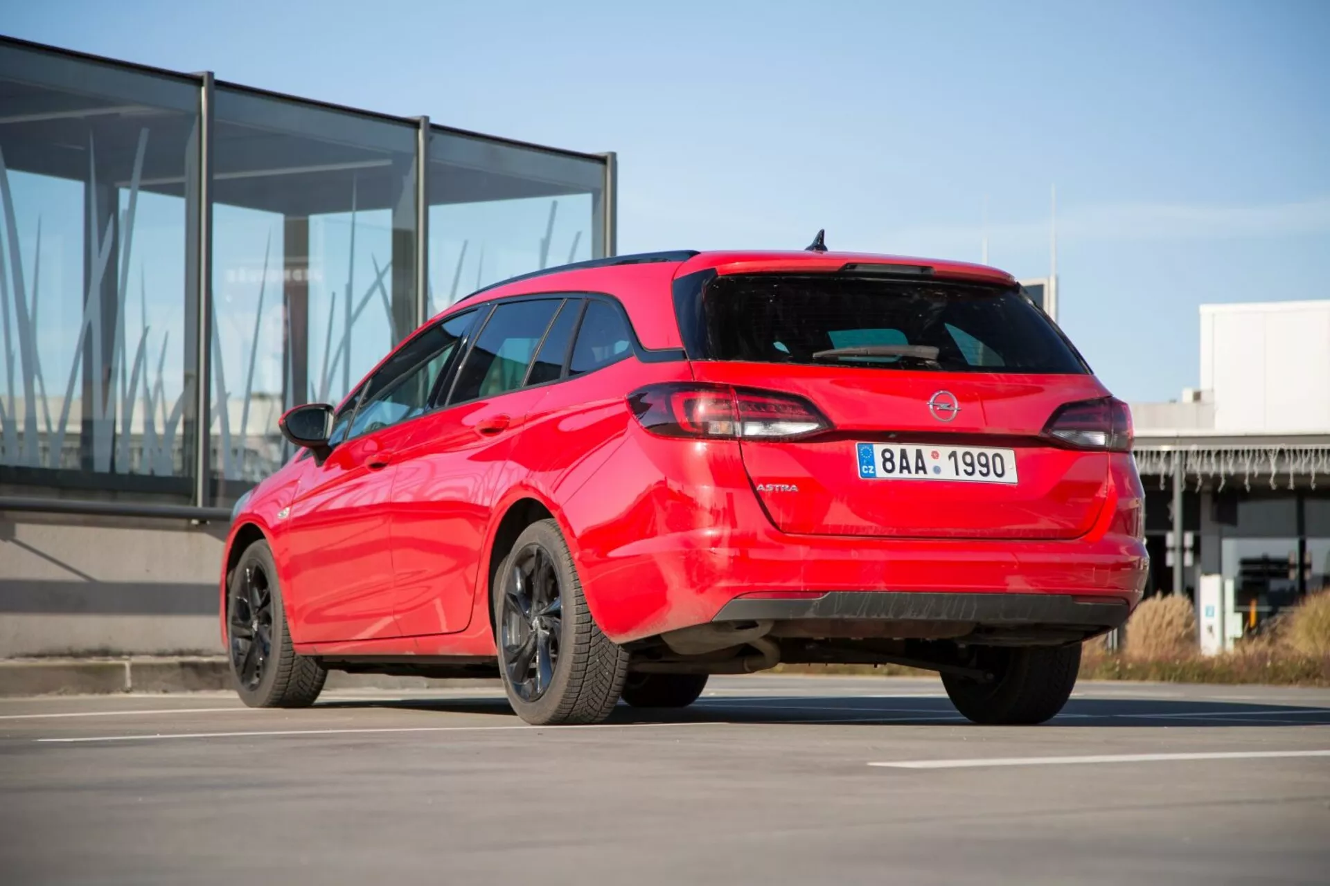 Rdeči hatchback Kia Ceed letnik 2009 prikazuje svoj športni zadnji tričetrtinski pogled na betonu z ozadjem modernega stavbe s stekleno fasado, ki izpostavlja značilen dizajn zadnjih luči, spojler na strehi, anteno v obliki morskega psa, lita platišča in morebitne senzorje za parkiranje.