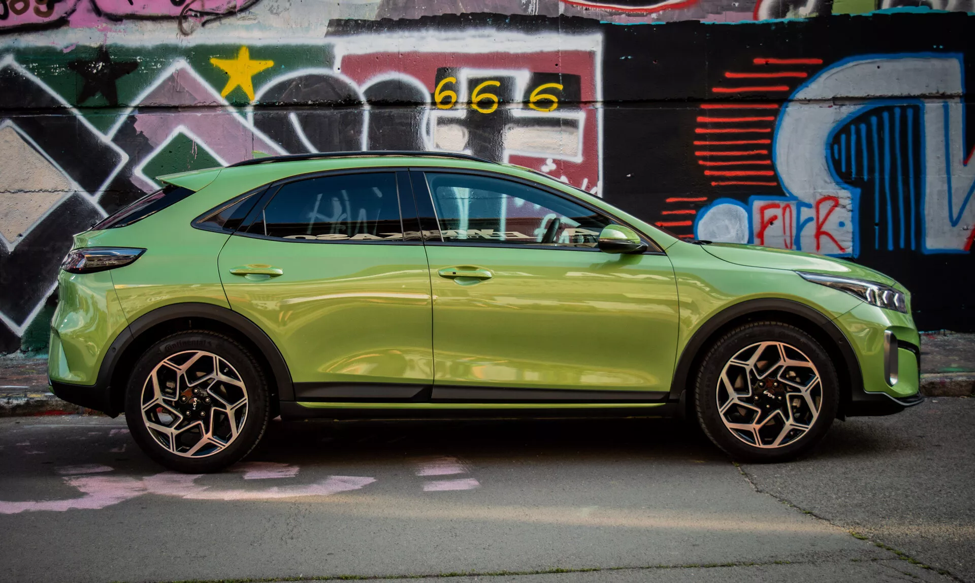 Kompaktni SUV Kia XCeed letnik 2022 v svetlo zeleni barvi, parkiran ob zidu pokritem z grafiti, razkazuje svojo elegantno zasnovo in napredne funkcije kot so brezključni vstop in vgrajeni smerniki v stranskih ogledalih.