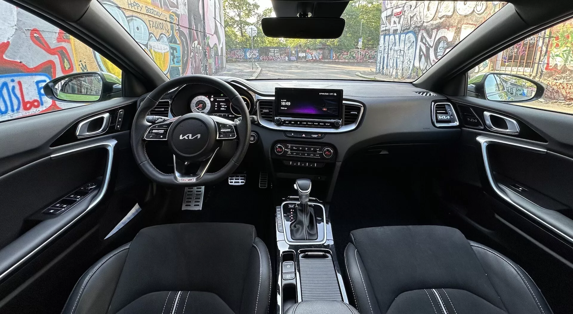 Notranjost Kia XCeed 2022, gledano iz zadnjih sedežev, prikazuje volan z nadzornimi gumbi, digitalni instrumentni zaslon, osrednji zaslon za zabavo in informacije, ročne nastavitve za klimo, sprednji sedeži, oblečeni v usnje, prestavna ročica v parkirnem položaju, in napredne funkcionalnosti, kot so nadzor voznih načinov in samodejno zatemnitev vzvratnega ogledala, vse v temni barvni shemi.