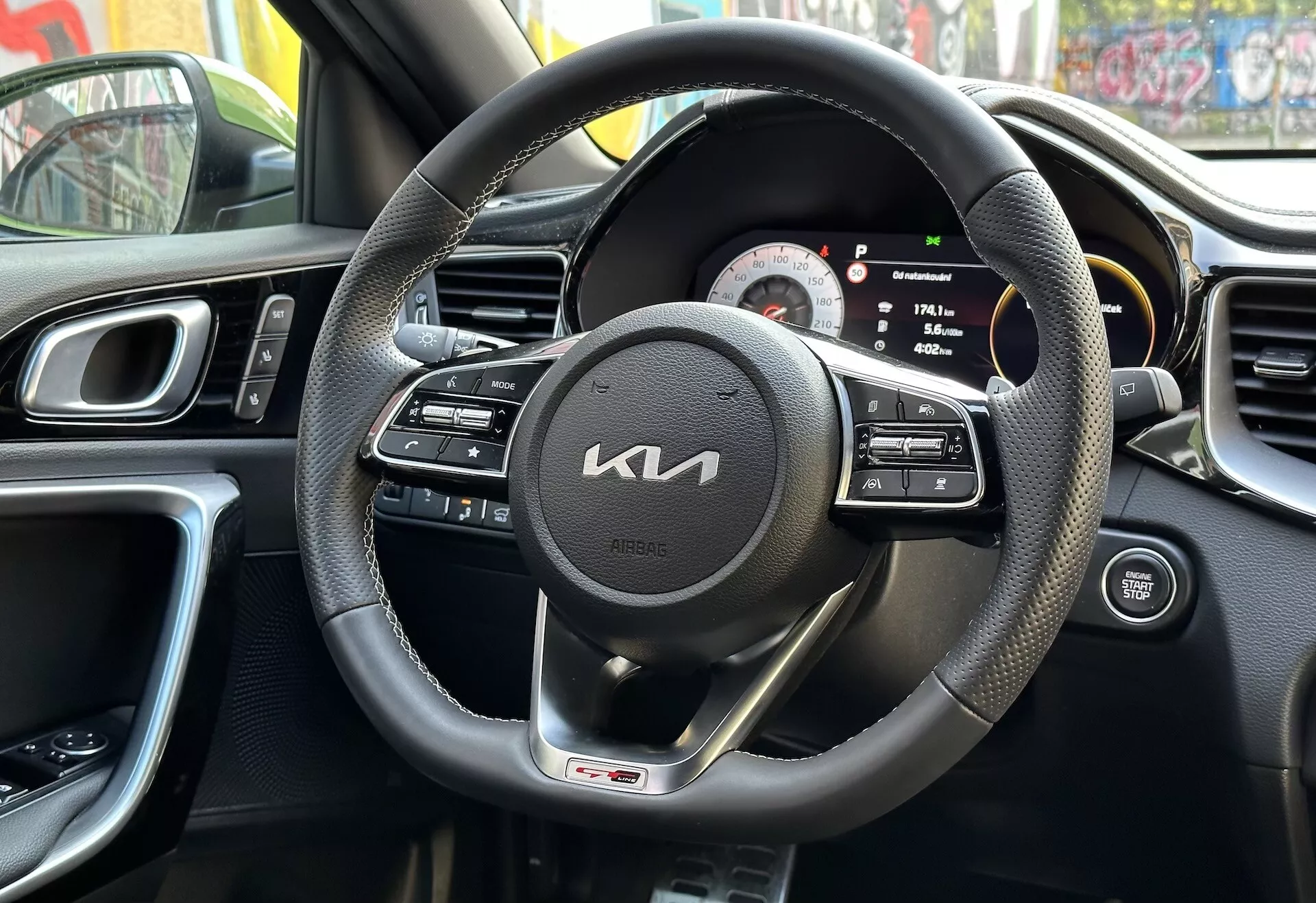 Pogled na notranjost Kia XCeed 2022 iz perspektive voznika z črnim usnjenim volanom z multifunkcijskimi gumbi in značko GT line, digitalno-analogni merilnik, del srednje konzole z zračniki, ergonomsko oblikovanje armaturne plošče in del vrata voznika z gumbi, odraža urbano okolje